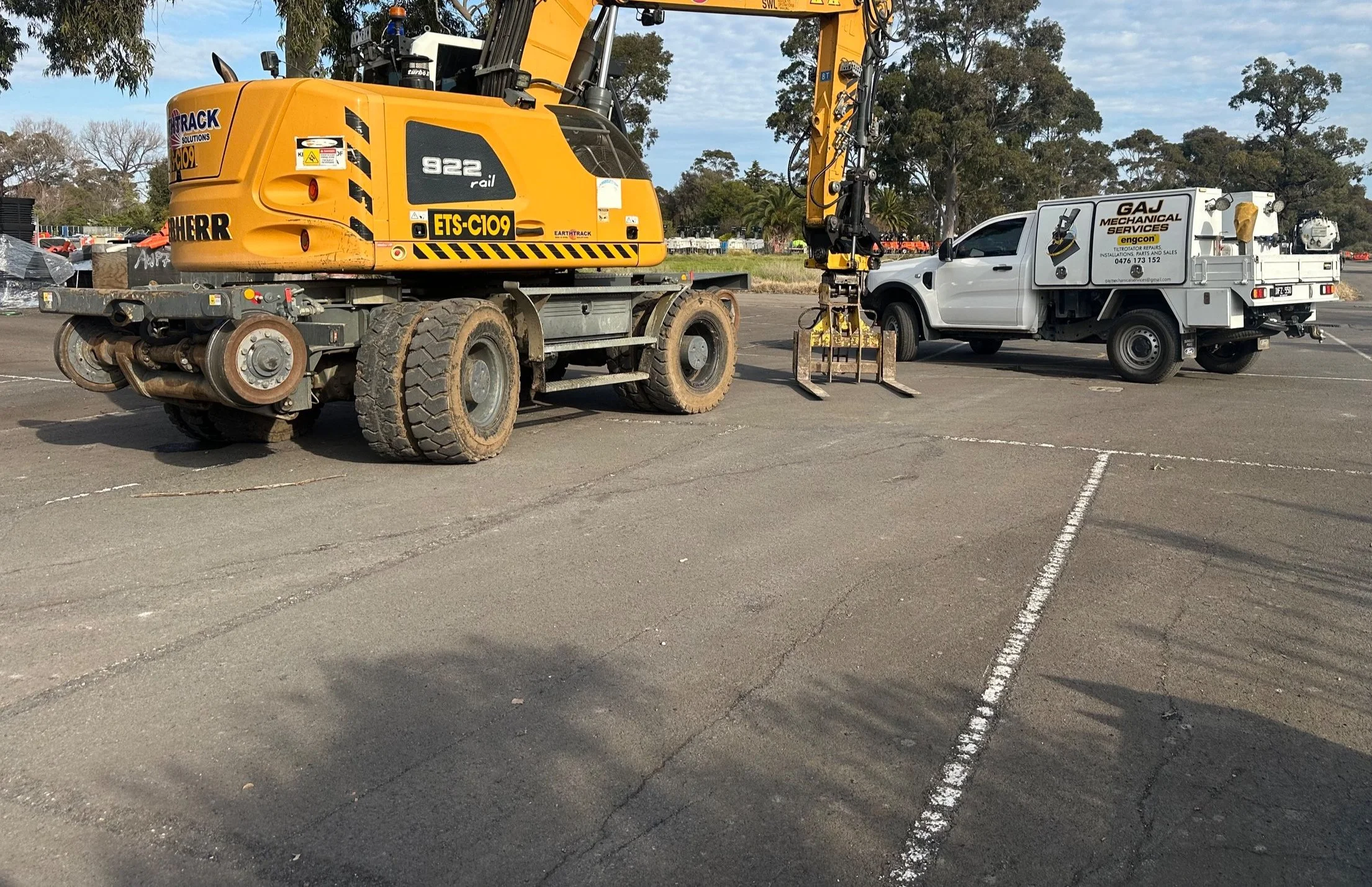 A construction site with a yellow liebherr hi-rail excavator and a white utility truck parked in a lot.