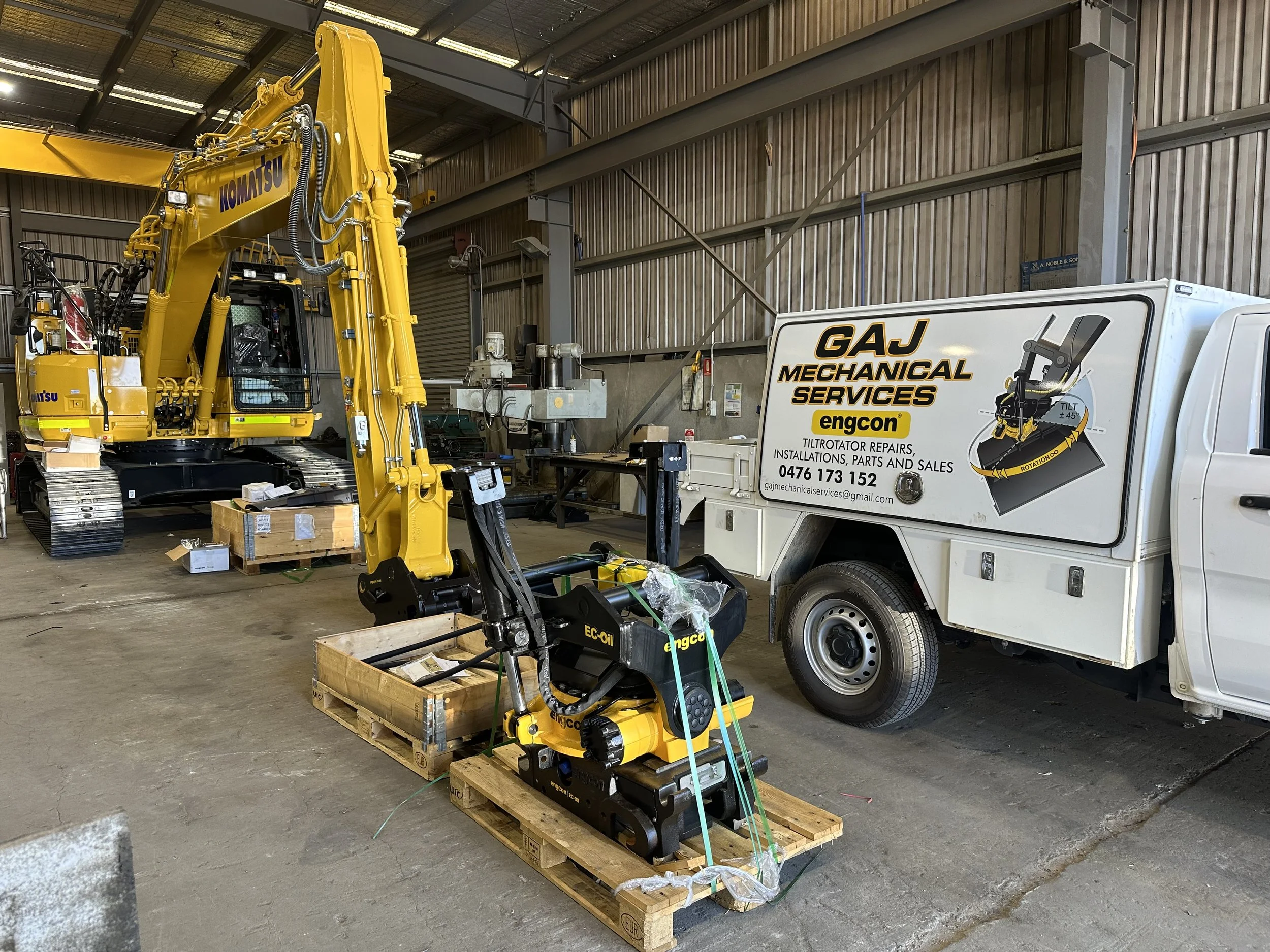 Industrial storage area with a yellow Komatsu excavator, a white service truck labeled GAJ Mechanical Services with contact info, and a small black and yellow engcon tiltrotator on a pallet.