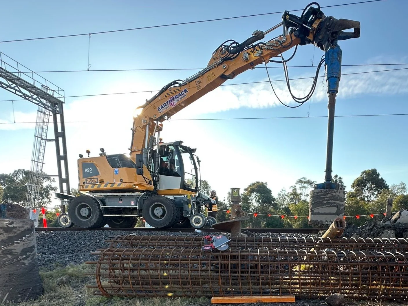 Construction site with an excavator drilling , surrounded by rebar and construction equipment, in the rail corridor, hi-rail excavator, under a clear sky.