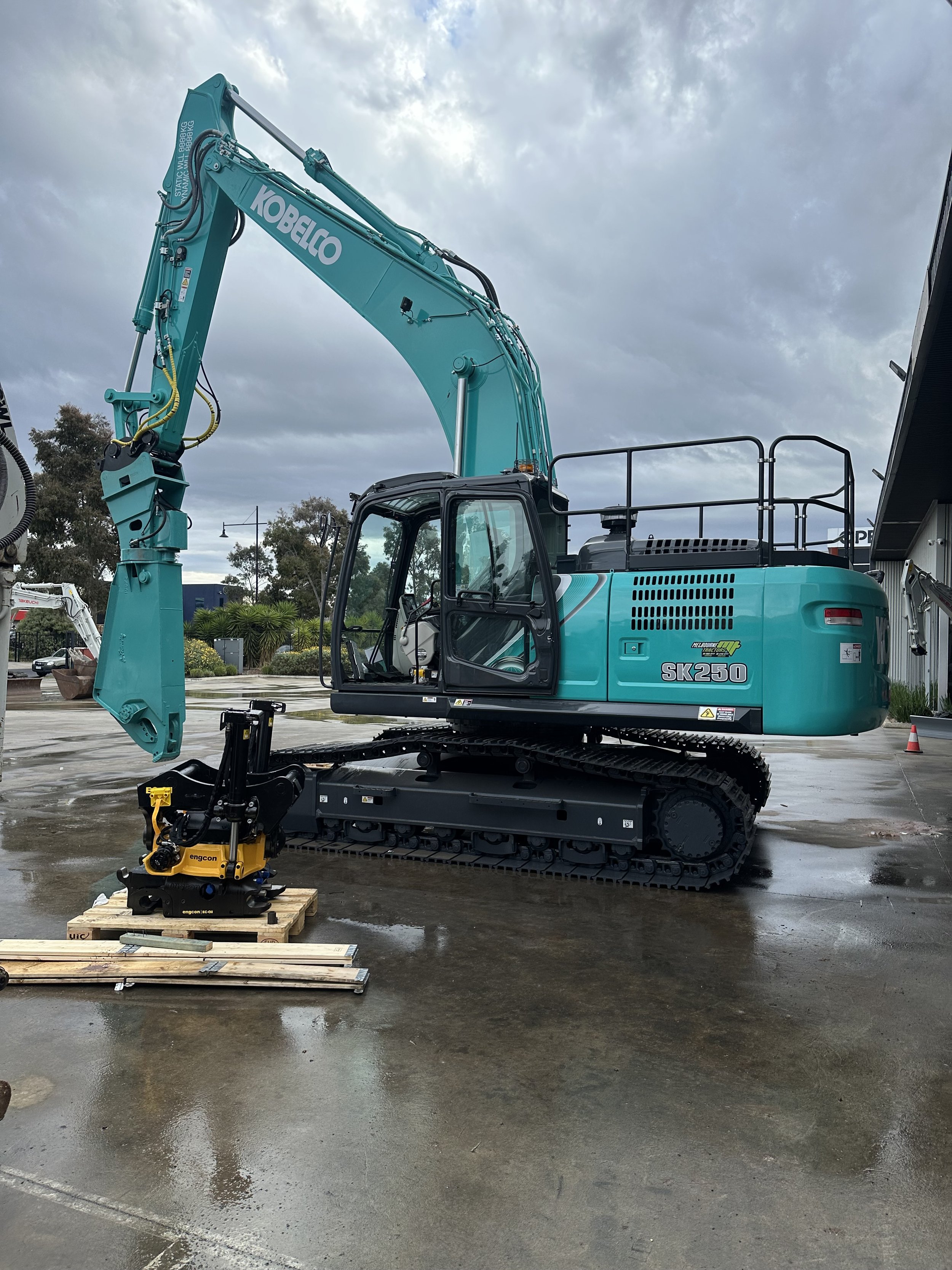 A teal and black Kobelco SK250 excavator with engcon tiltrotator and shear parked on a wet concrete surface in an outdoor lot, with a cloudy sky and some trees and other construction equipment in the background.
