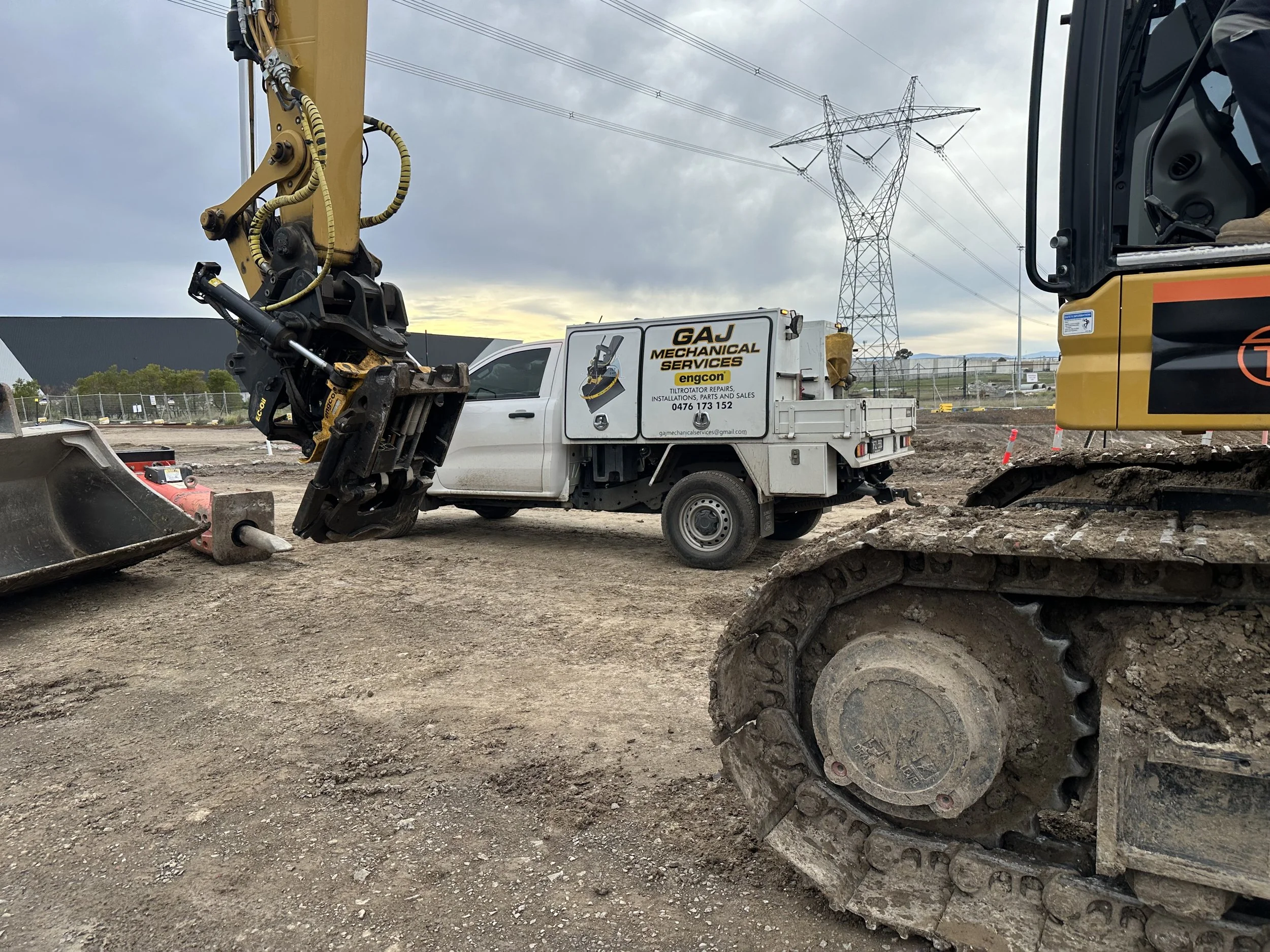 Construction site with a large excavator and tiltrotator, a utility truck with company signage, and power lines in the background.