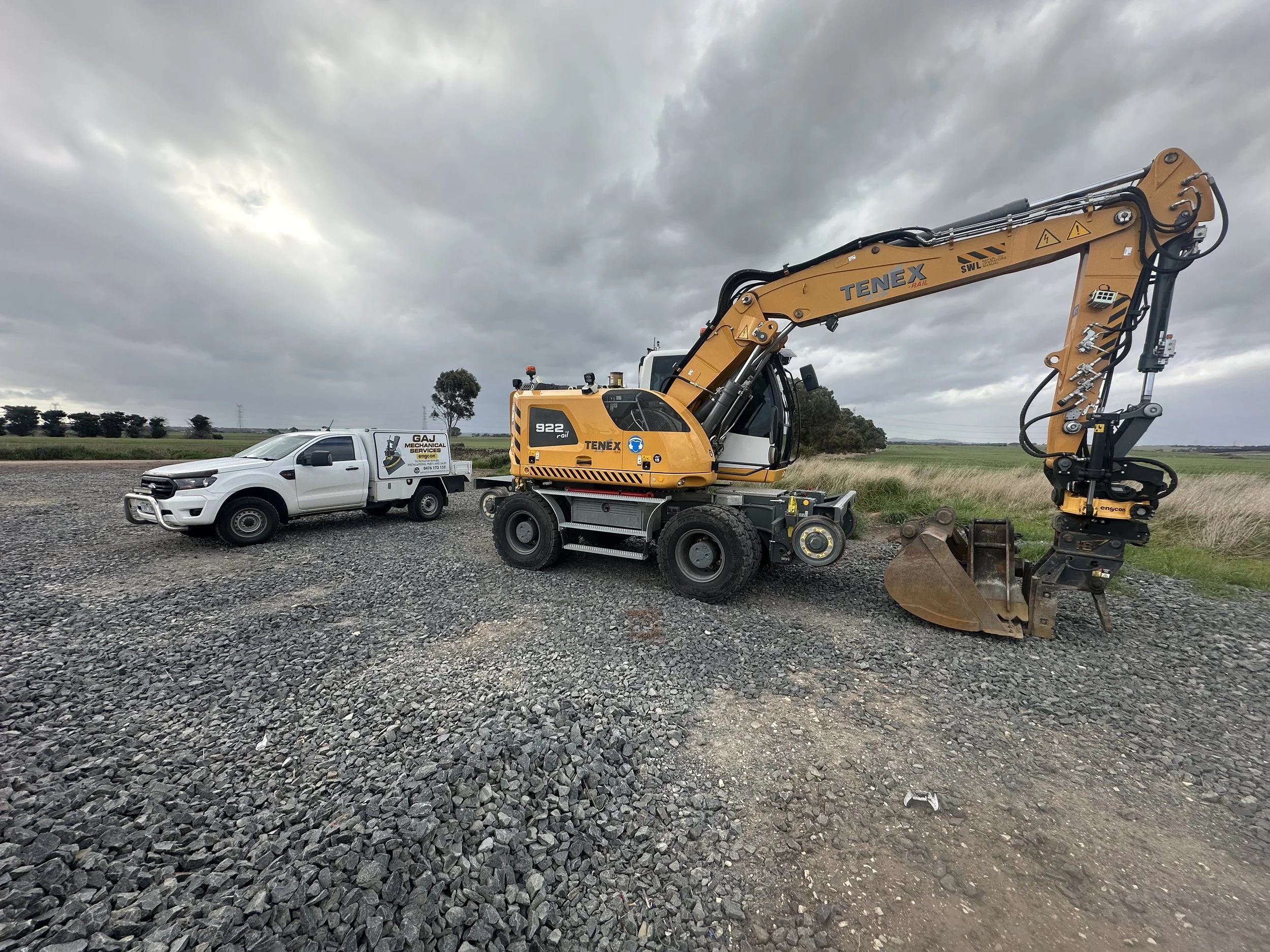 A white utility truck and a large yellow excavator parked on gravel, with open countryside and cloudy sky in the background in the victorian rail corridor near Melbourne