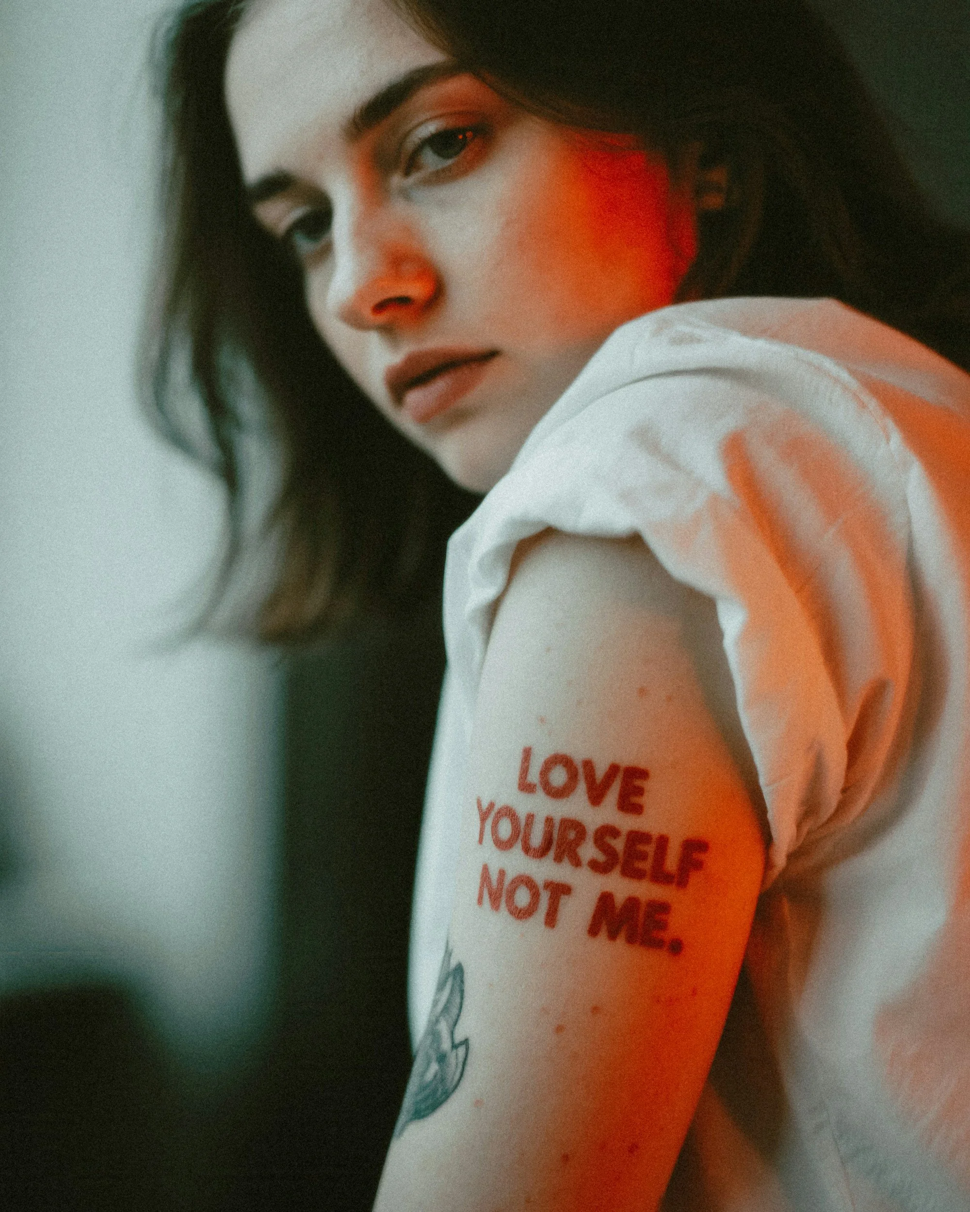Close-up of a woman with dark hair, looking at the camera with a serious expression, wearing a white shirt with a tattoo on her arm that reads 'Love yourself not me' in red letters.