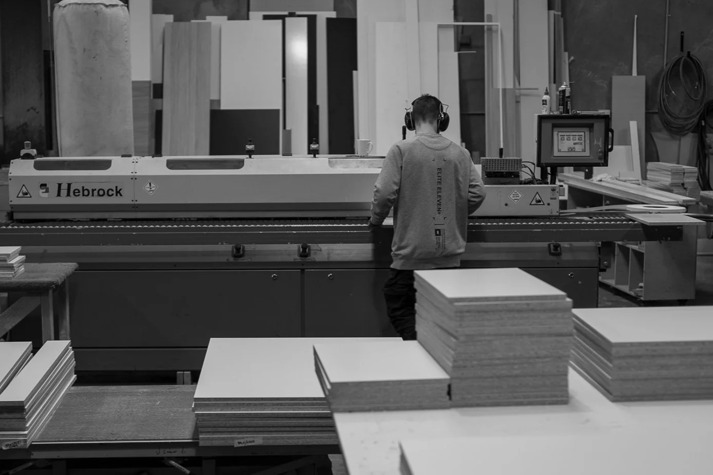 Brody wearing headphones operating an edging machine in our woodworking workshop with stacks of cut cabinet materials in the foreground.