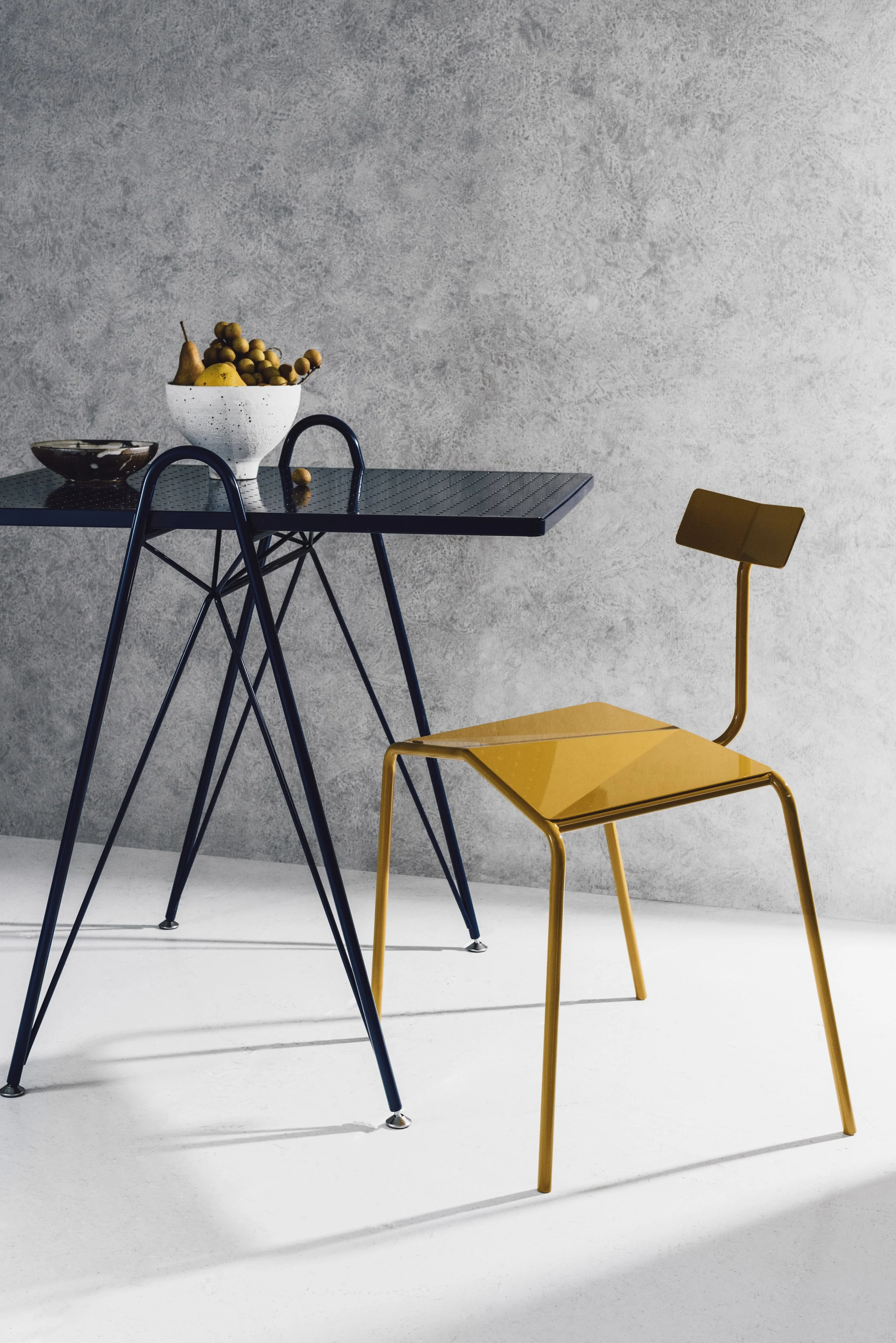 A modern black dining table with blue metal legs, a yellow metal chair, and a gray textured wall in the background. On the table, there is a white bowl of fruit, including grapes, pears, and a banana, along with a small black dish.