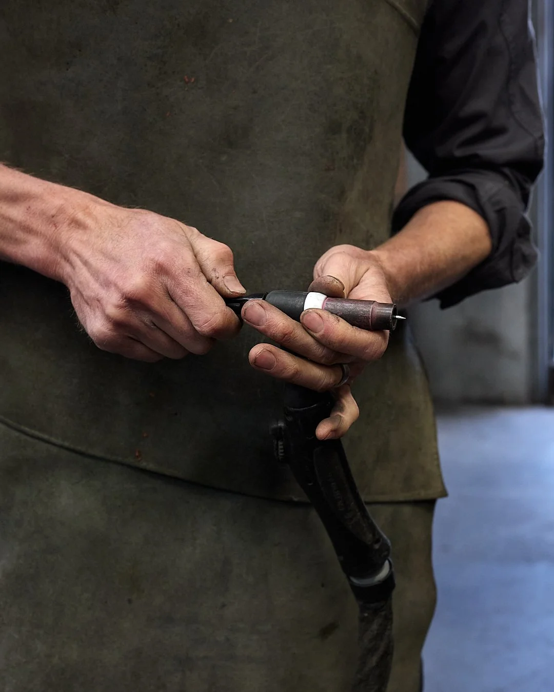 Close-up of Dan's hands, holding a welding torch, wearing a dark apron, working in the workshop.
