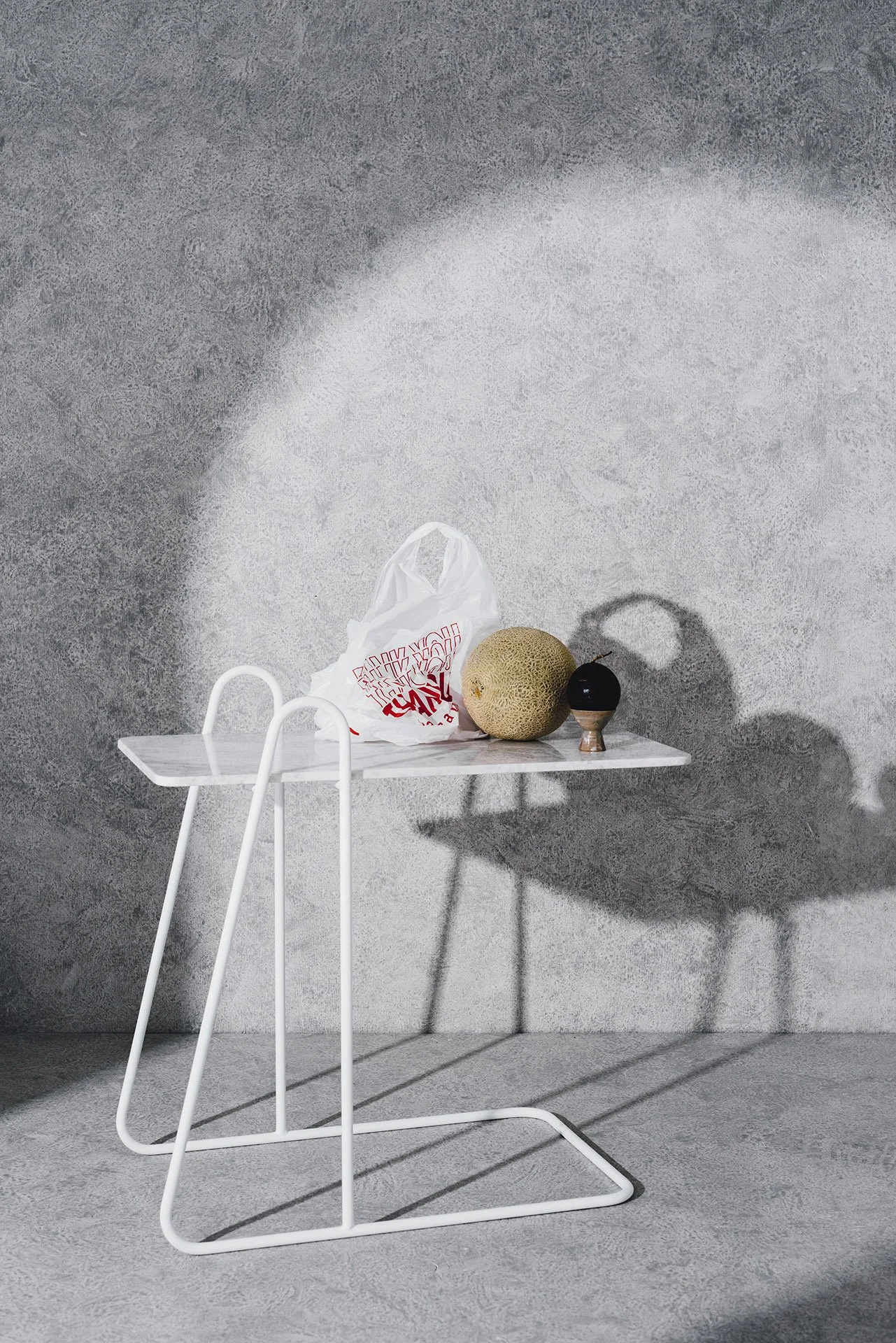 A minimalist scene with a custom white metal side table holding a plastic bag, a cantaloupe, and a small black vase with a shadow cast on a textured gray wall.