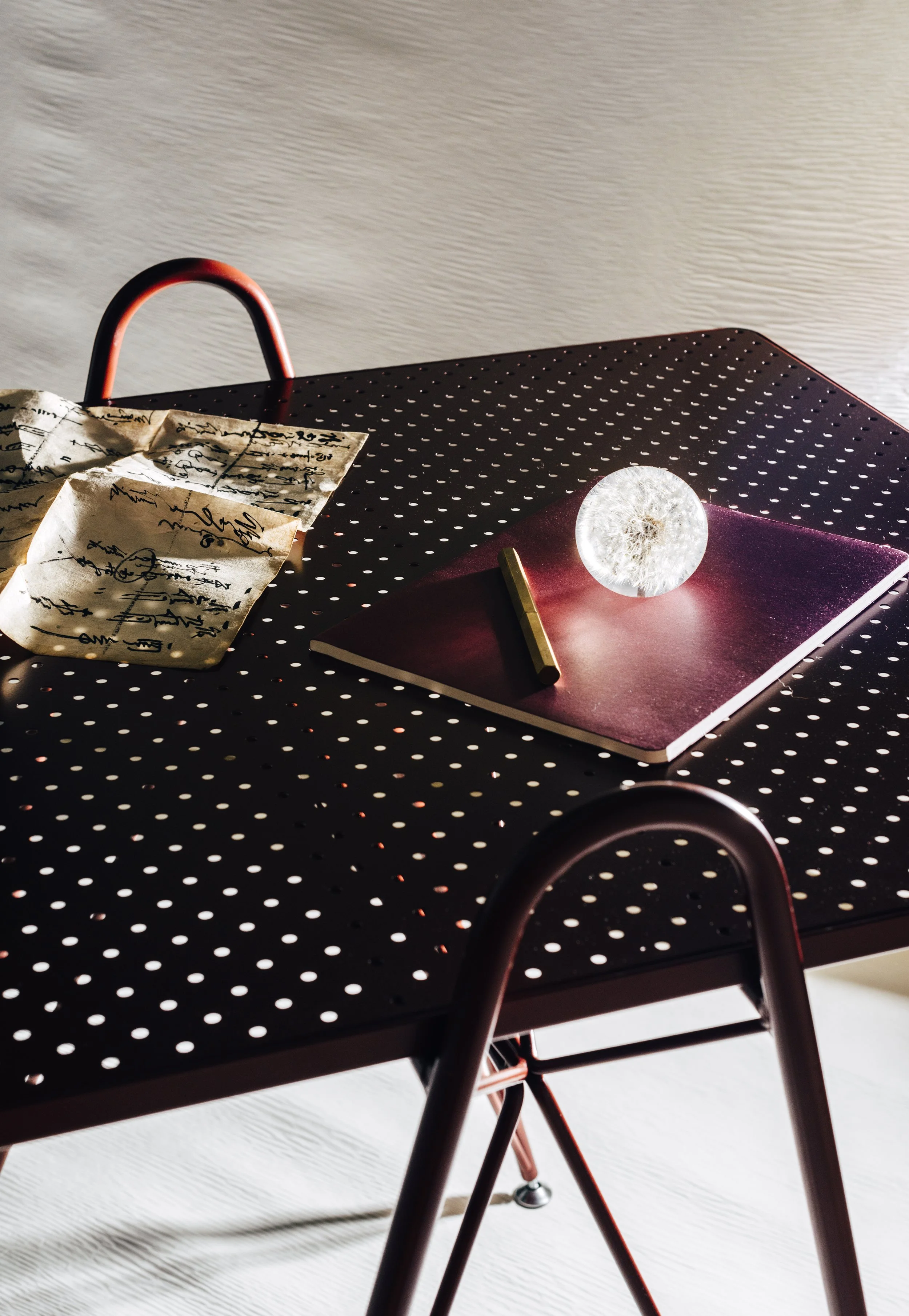 Custom metal table with scattered crumpled paper, a red notebook, a dandelion in a glass dome, and a gold pen on a polka dot metal table, with sunlight casting shadows.