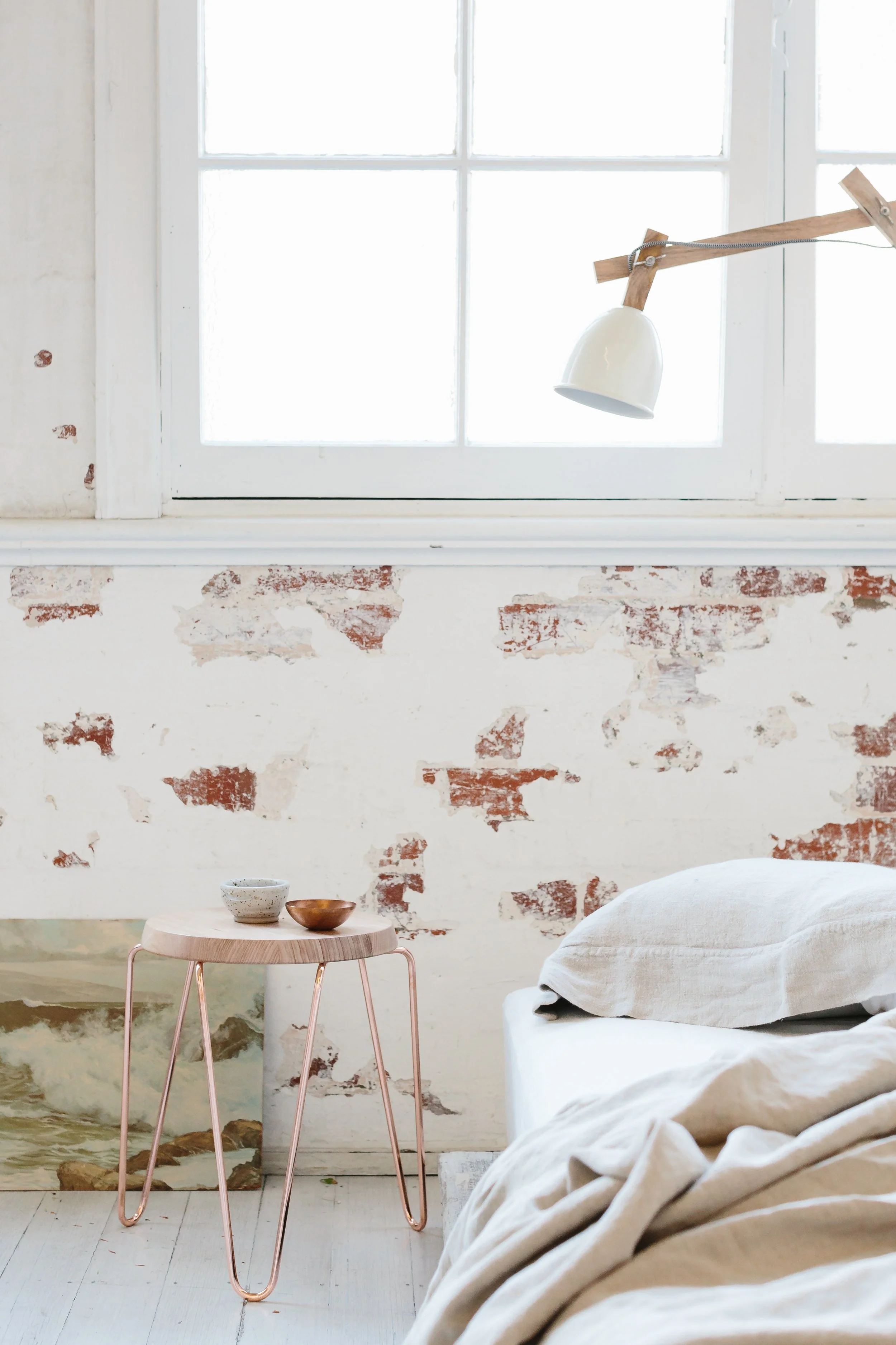 A minimalist bedroom with a white wall featuring exposed red bricks, a window, a small round side table with two bowls, an unmade bed with beige linen, and a wall-mounted adjustable lamp.