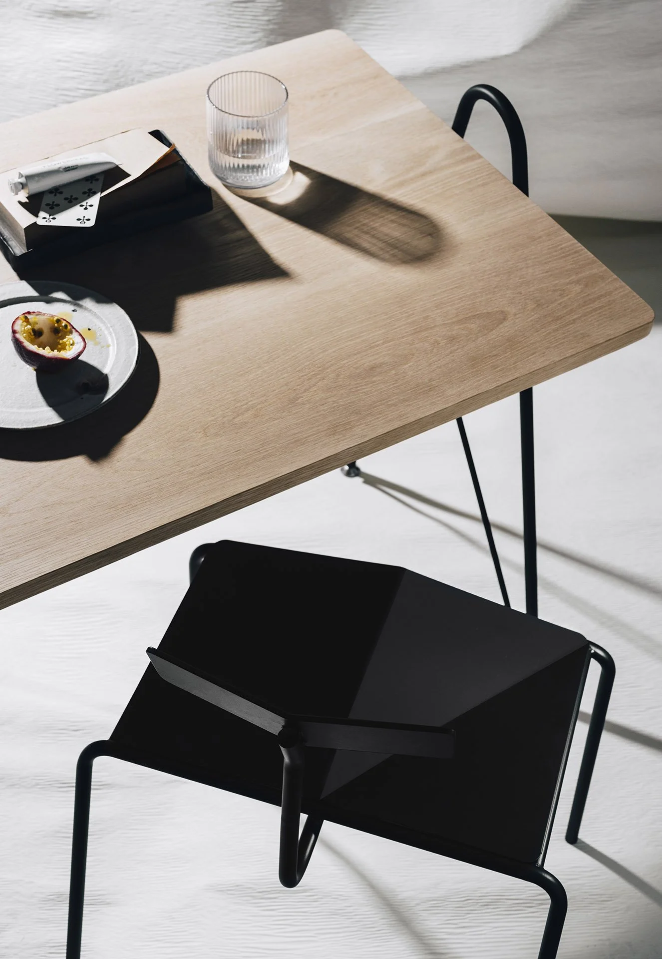 A custom wooden table with a half-cut passion fruit on a white plate, a glass of water, an open notebook with a pen, and playing cards inside a box, with shadows cast on the table and chair.