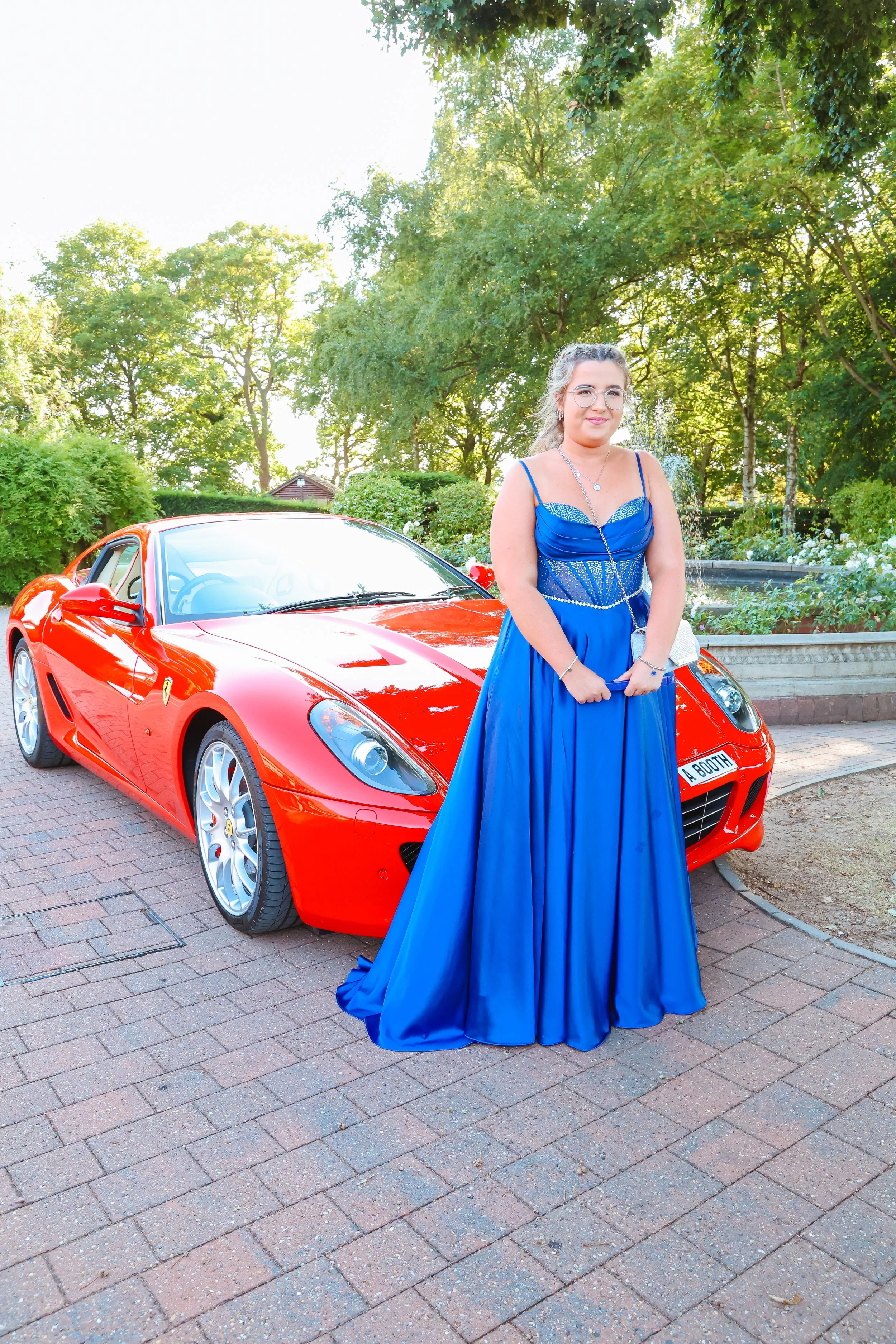 A young woman in a royal blue evening gown standing next to a red sports car on a brick-paved path with green trees and shrubbery in the background.