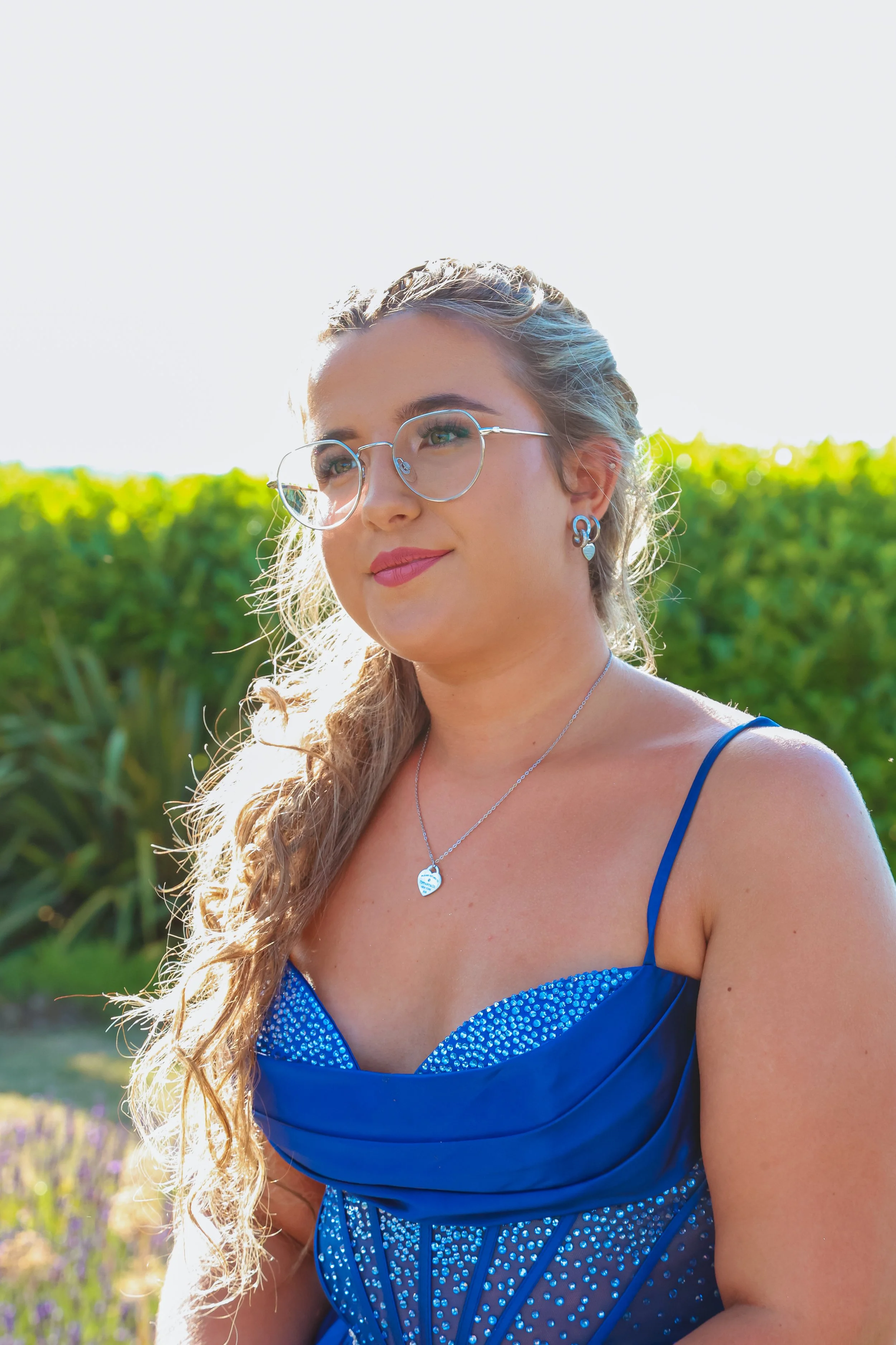 A woman with long, curly hair in a blue dress with rhinestones, wearing glasses, earrings, and a necklace, standing outdoors near greenery.