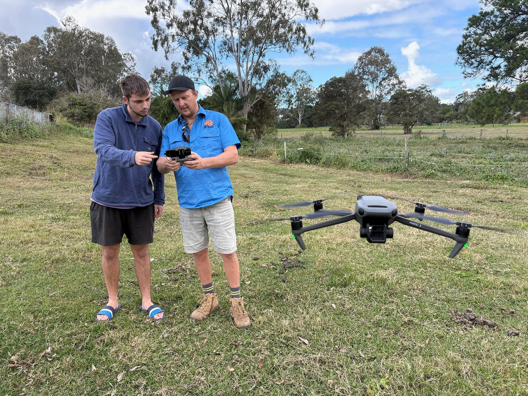 Two men standing outdoors in a grassy field, operating a remote-controlled drone flying nearby. The man on the left is wearing a blue jacket and black shorts, while the man on the right is wearing a blue short-sleeved shirt, beige shorts, and a black
