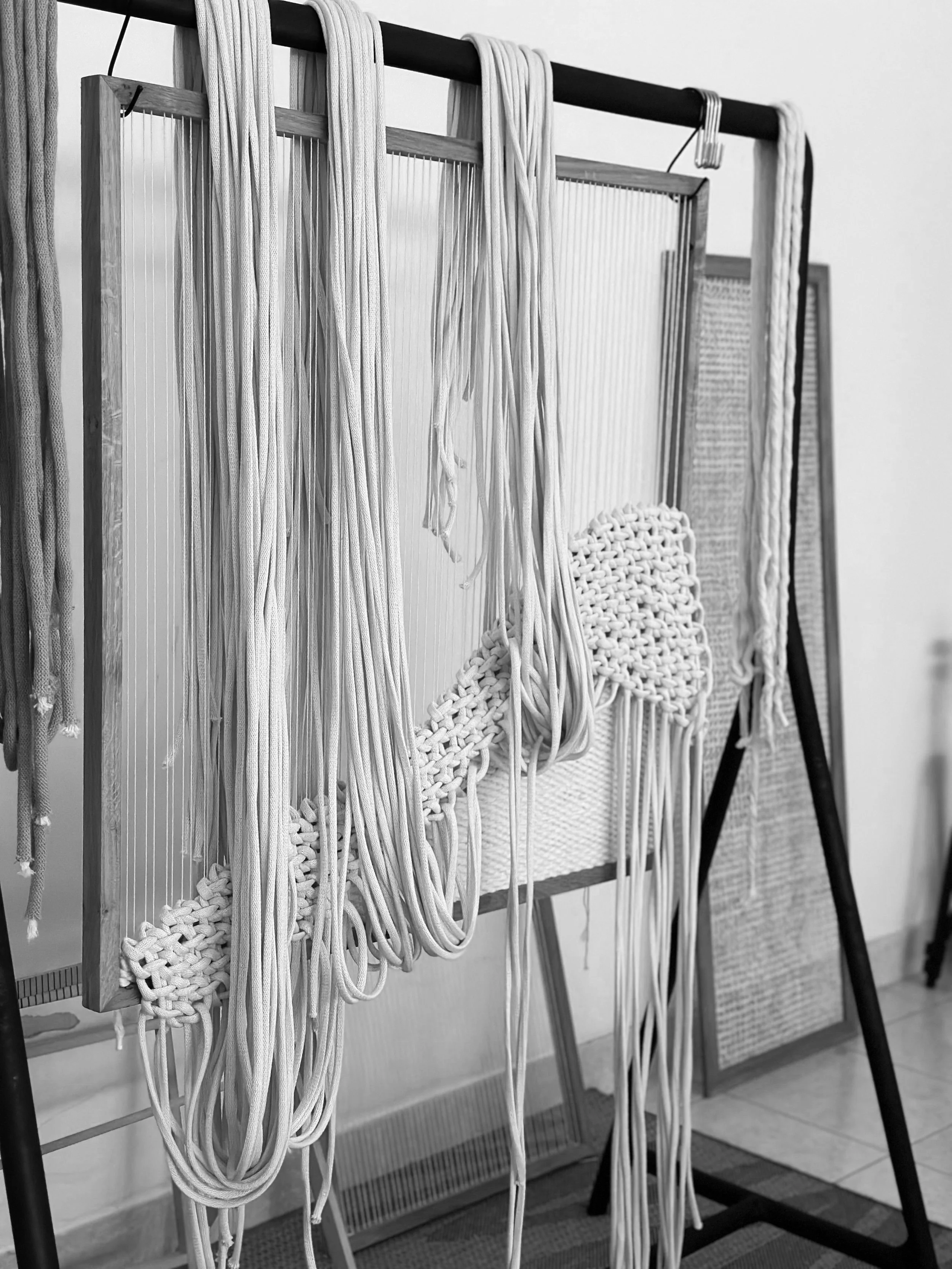 A display of various lengths and thicknesses of white macrame cords and ropes hanging on a black metal stand with a wooden frame behind them, used for creating macrame crafts.