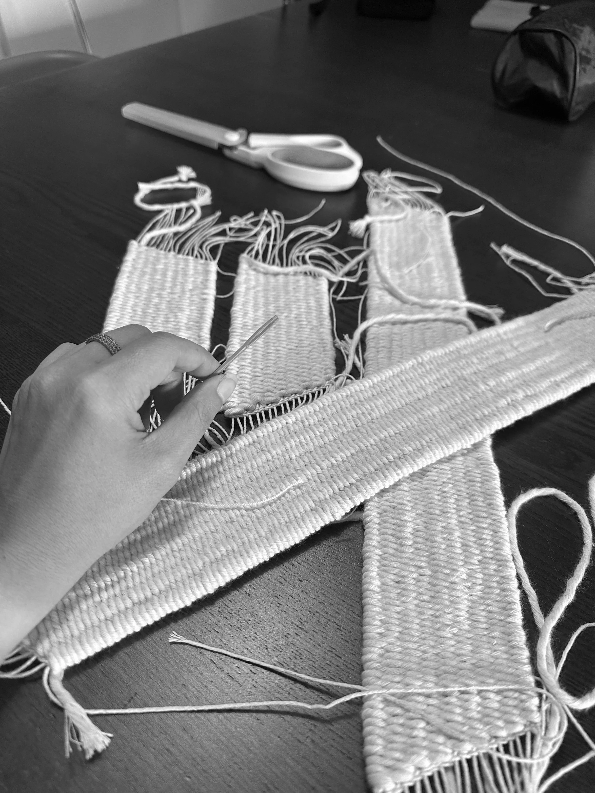 Close-up of a person's hand sewing a woven fabric with a crochet hook on a table, surrounded by partially completed woven strips, scissors, and sewing tools.