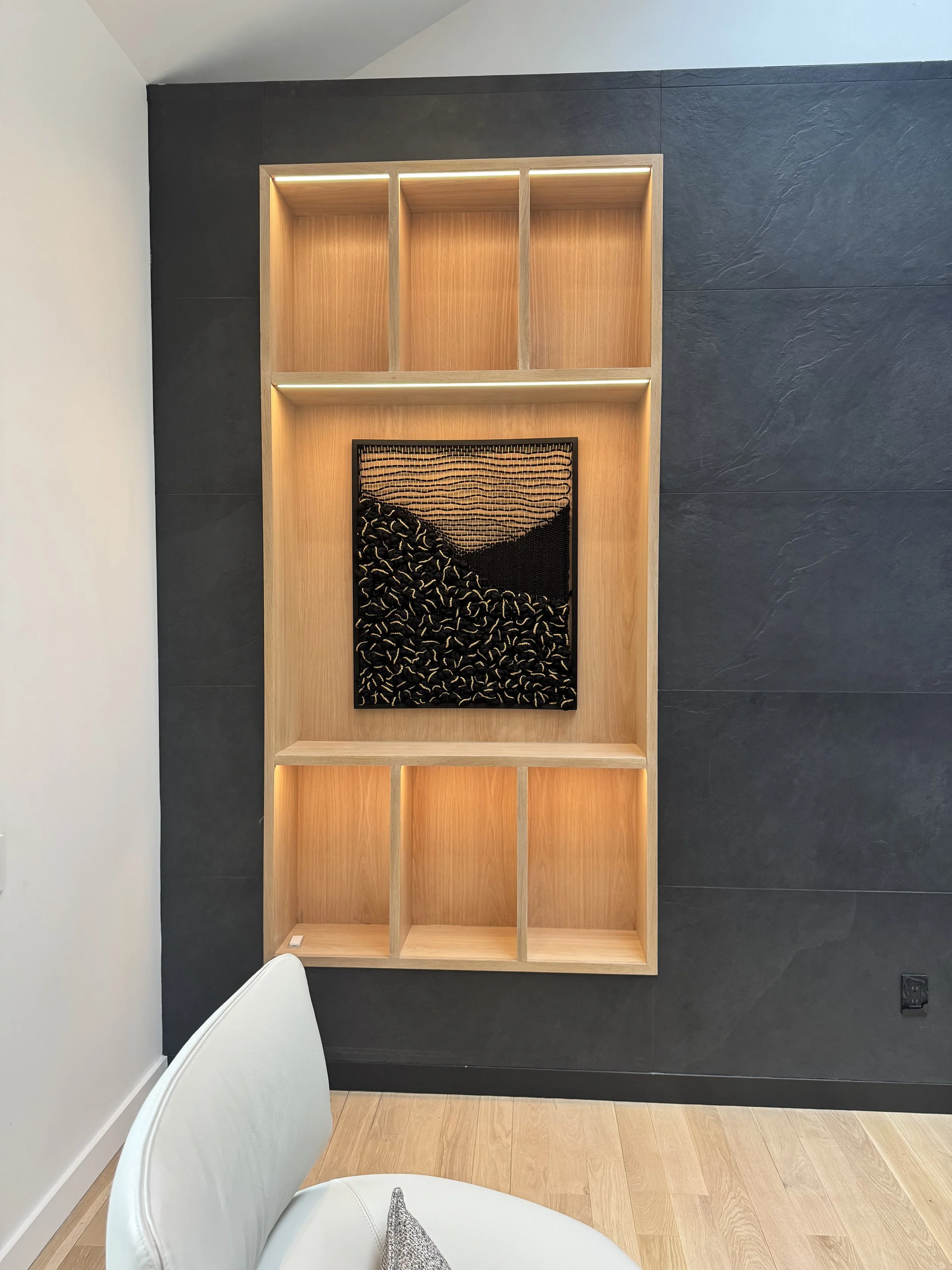 A modern wall with a wooden shelf and cubbies, containing a black woven art piece by textile artist Ana Salazar Atelier, against a dark textured wall and a light wood floor, with a white chair in the foreground.