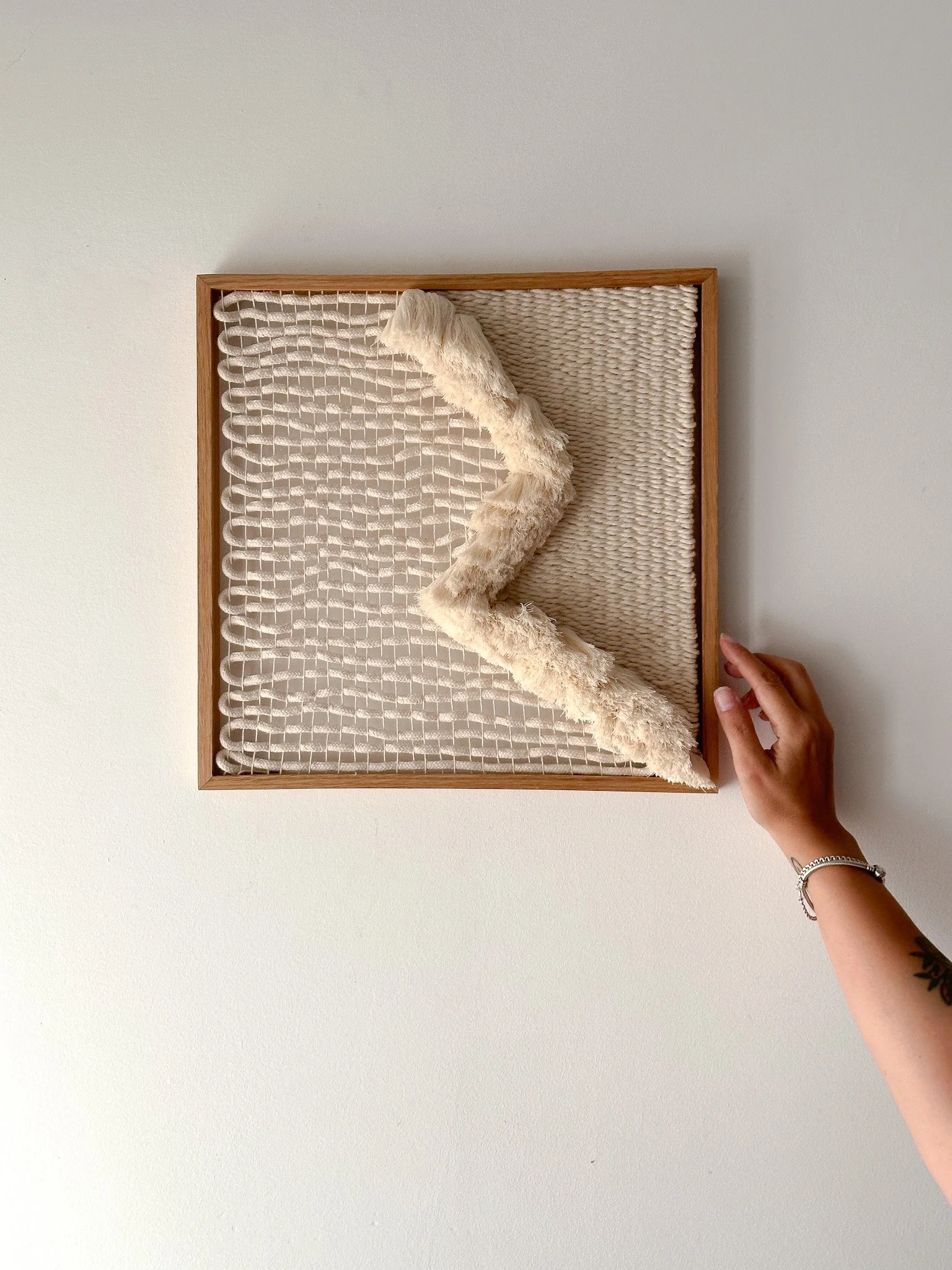 Person's hand holding the edge of a framed piece of textile art with cream, textured fabric and a wooden border, mounted on a plain white wall.