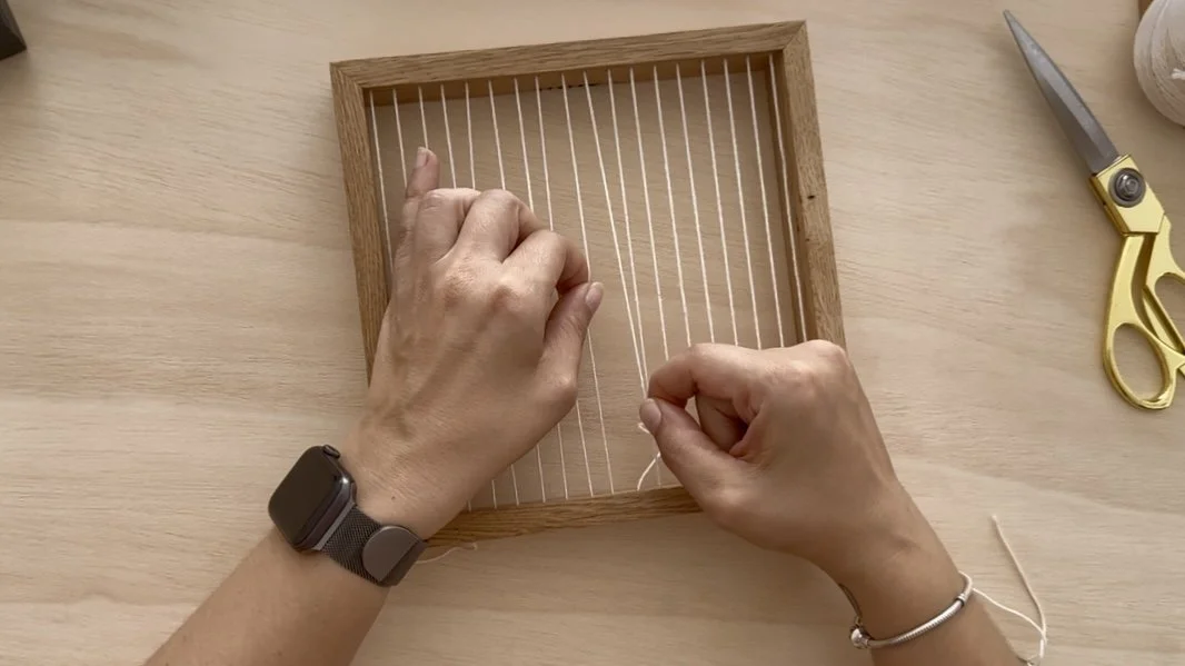 Hands weaving white natural cotton on a integrated weaving frame in oak with scissors and yarn balls nearby | Ana Salazar Atelier