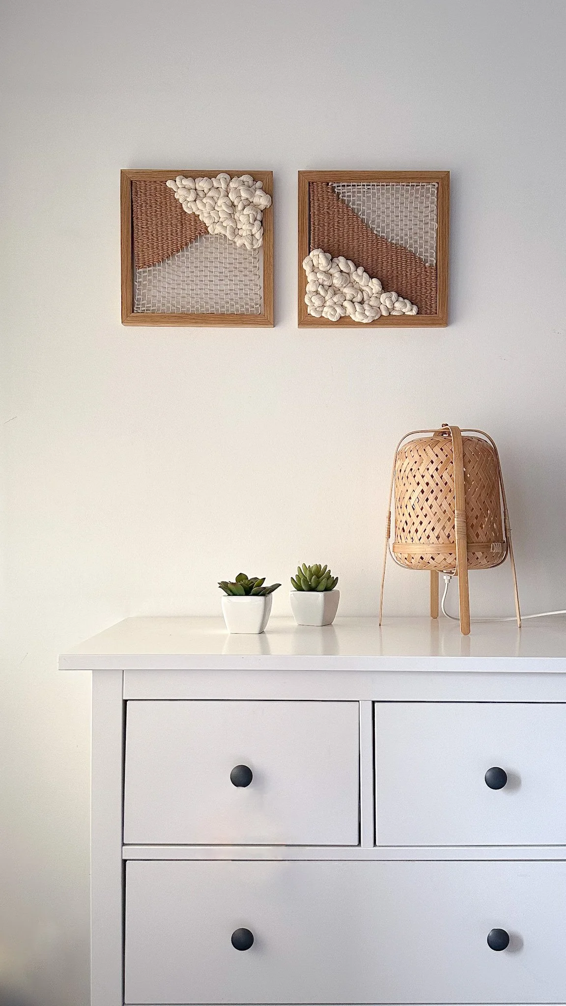 Minimalist interior decor with two framed textile art in earthy tones hung on a white wall above a white dresser with three drawers, adorned with two small potted succulents and a woven lamp | Ana Salazar Atelier