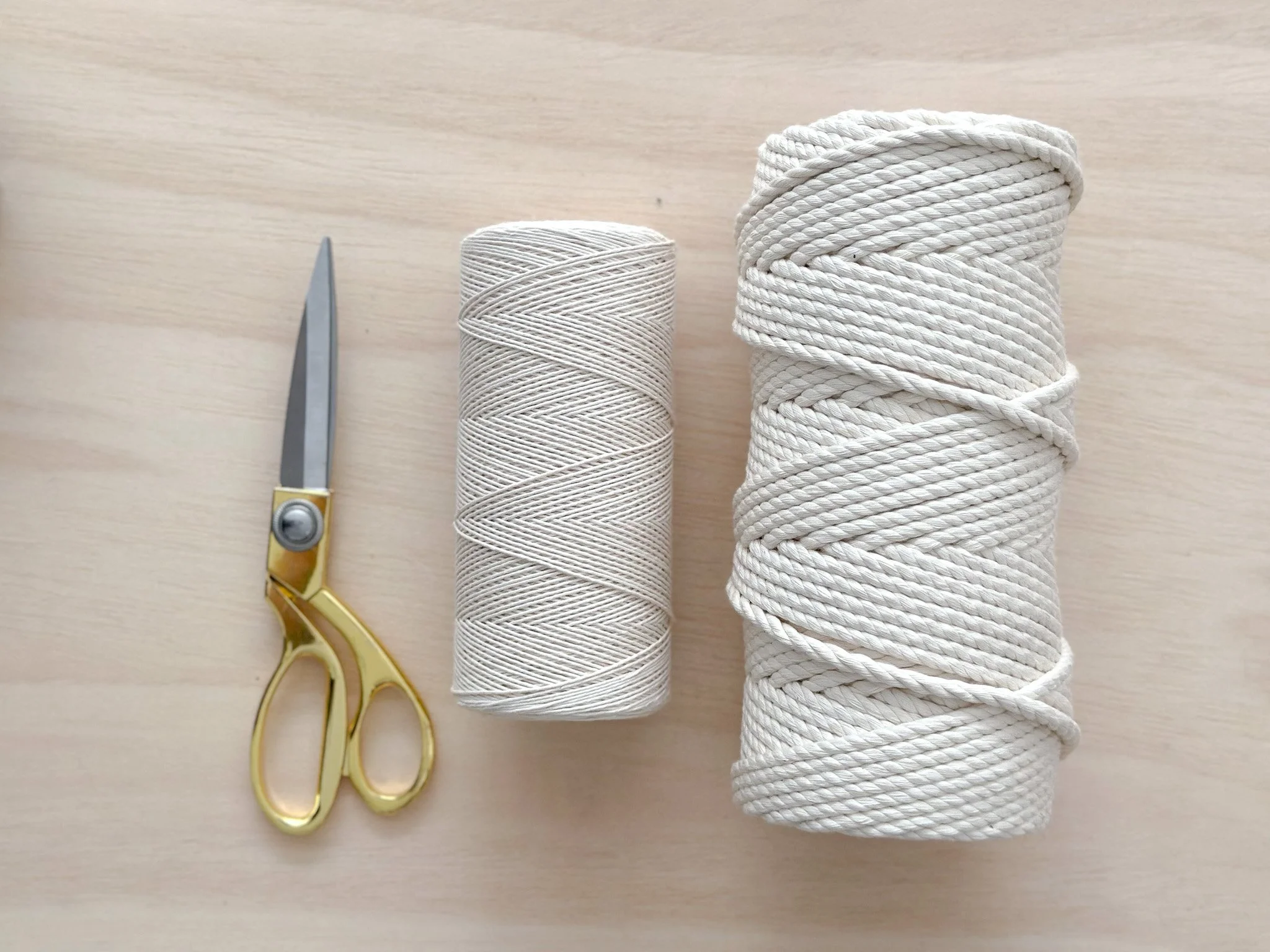 A pair of small scissors with gold handles, a spool of beige thread, and a larger spool of white twisted rope on a light wooden surface.