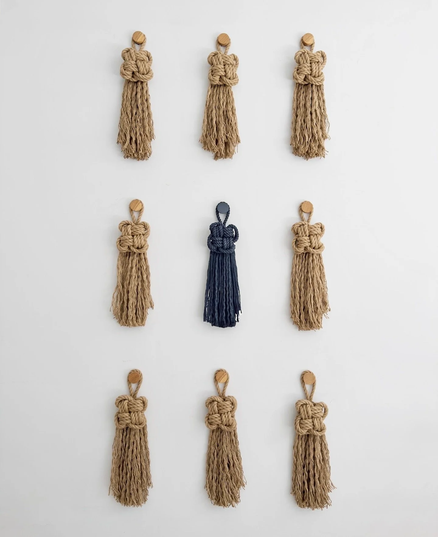 Nine large scale decorative knots with fringes, hanging on a white wall, one in the center is black, the rest are jute | Ana Salazar Atelier