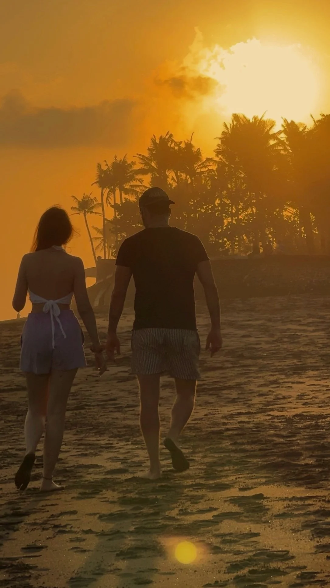Una coppia cammina sulla spiaggia al tramonto, con palme sullo sfondo e un cielo arancione.
