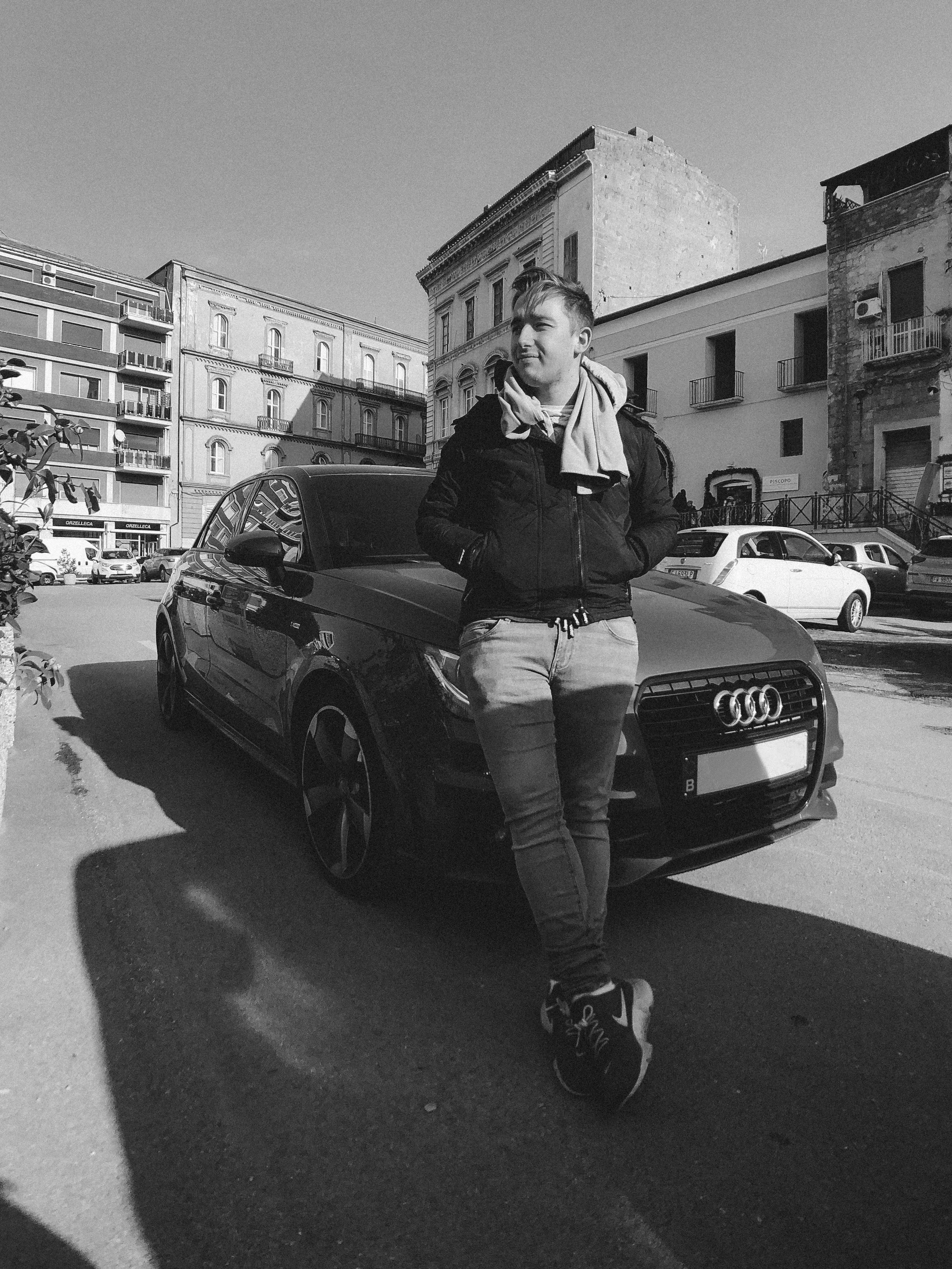Jeune homme debout devant une voiture noire Audi, portant un manteau noir, un hoodie, un pantalon beige clair et des baskets, dans une ville avec plusieurs bâtiments résidentiels et des voitures en arrière-plan.