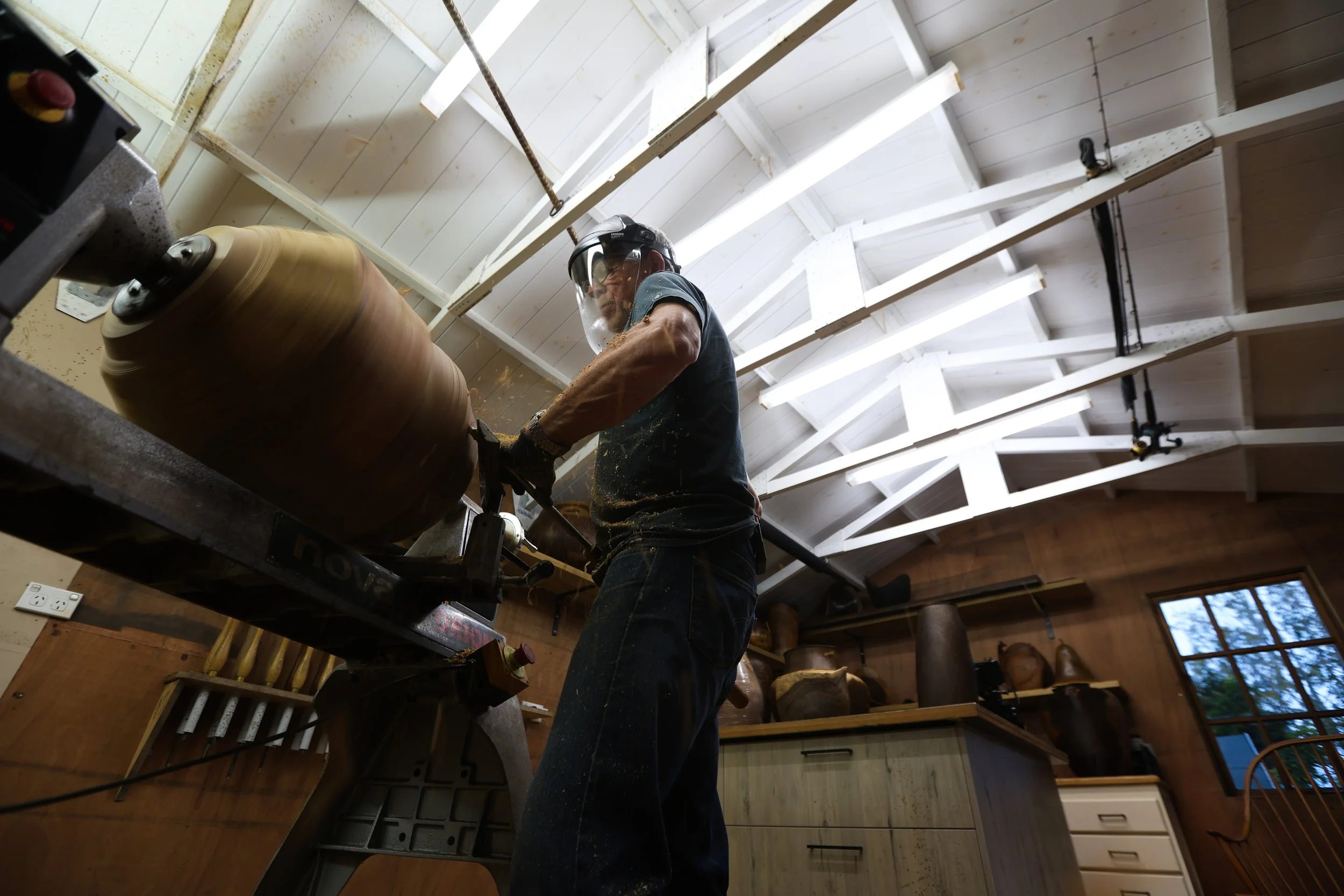 A person wearing safety gear, including a face shield, is operating a wood lathe to turn a piece of wood. The woodworking shop has wooden walls, tools, and supplies visible in the background.