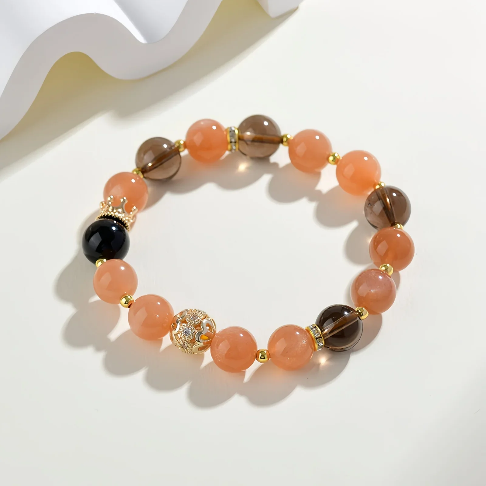 A crystal bracelet made of natural sunstone, natural black obsidian and natural smoky quartz with gold charms on a cream-coloured surface with a white, wave-like structure beside