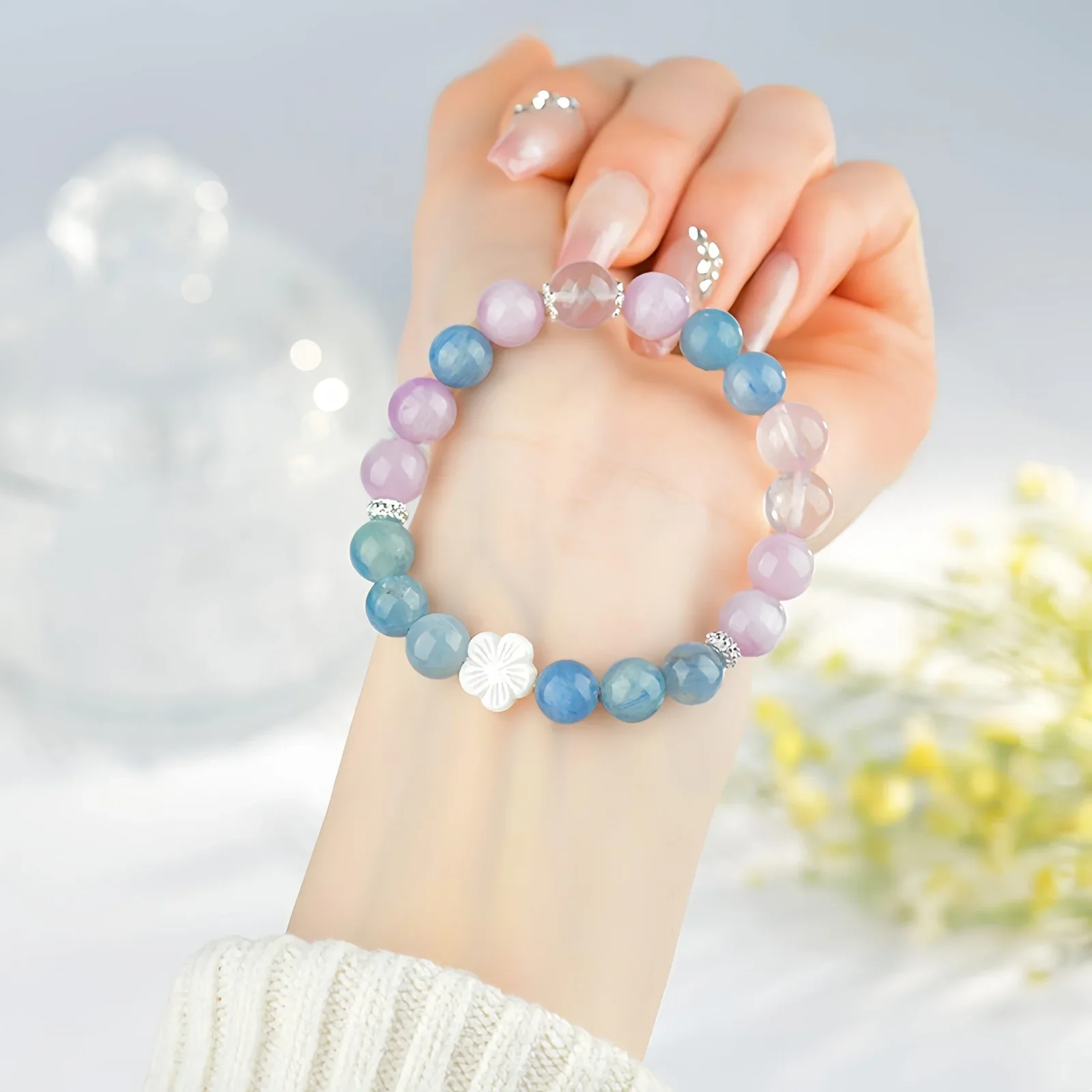 A crystal bracelet made of natural rose quartz, natural aquamarine and one fleur glass bead resting on a wrist with some yellow flowers and glassware in the background