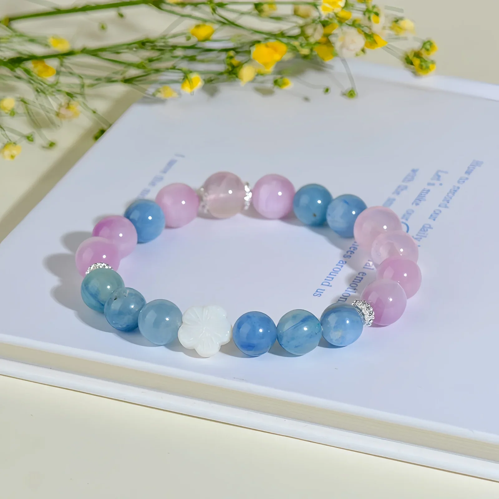A crystal bracelet made of natural rose quartz, natural aquamarine and one fleur glass bead resting against a book on a cream-coloured surface with some small yellow flowers in the background