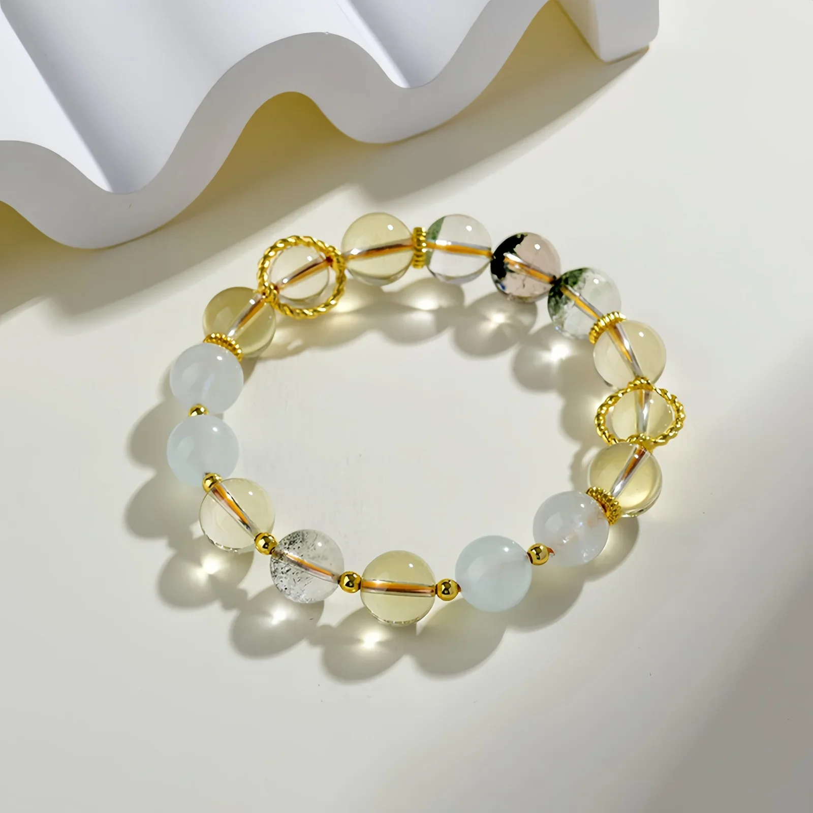 A crystal bracelet made of natural white quartz, natural green phantom quartz and natural citrine on a cream-coloured surface with a white, wave-like structure beside it