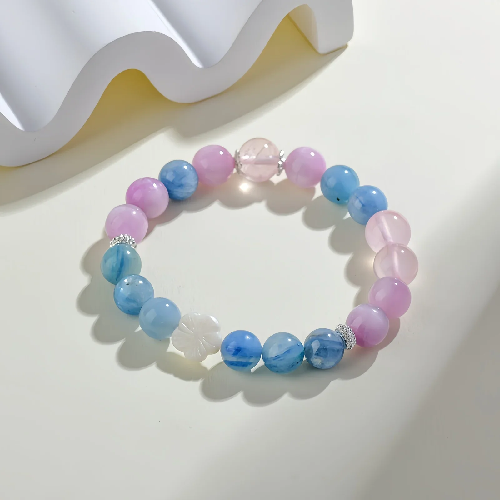 A crystal bracelet made of natural rose quartz, natural aquamarine and one fleur glass bead on a cream-coloured surface with a white, wave-like structure beside it