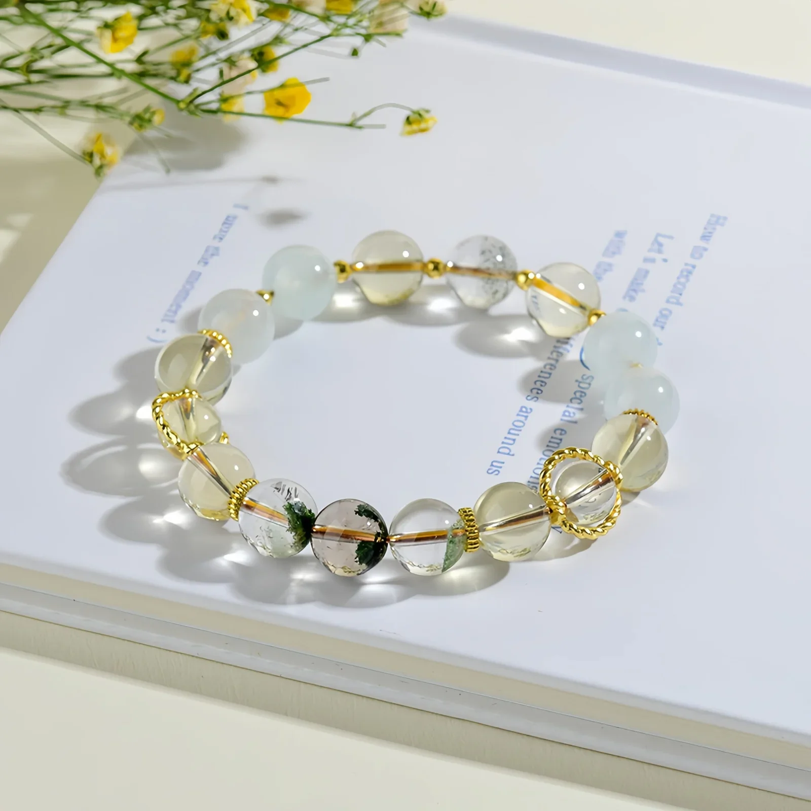A crystal bracelet made of natural white quartz, natural green phantom quartz and natural citrine resting on a book on a cream-coloured surface with tiny yellow flowers in the background