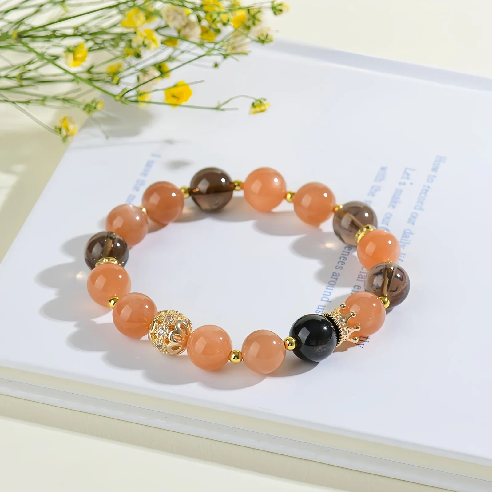 A crystal bracelet made of natural sunstone, natural black obsidian and natural smoky quartz with gold charms on a book with some small yellow flowers beside it