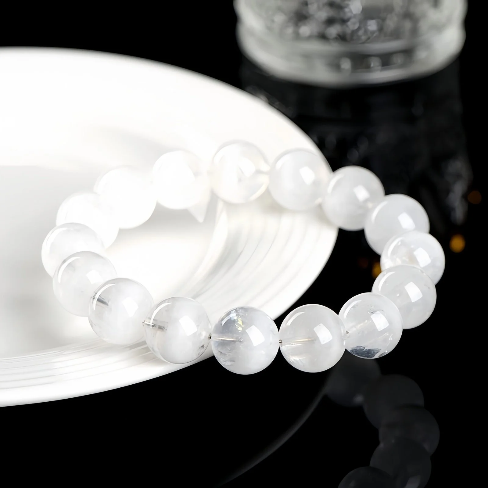 A natural white phantom quartz bracelet on a white plate against a glossy black surface beside a glass of water