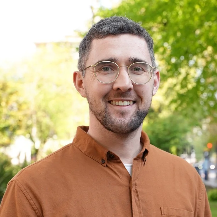 A young man with glasses and a beard smiling outdoors in front of green trees.