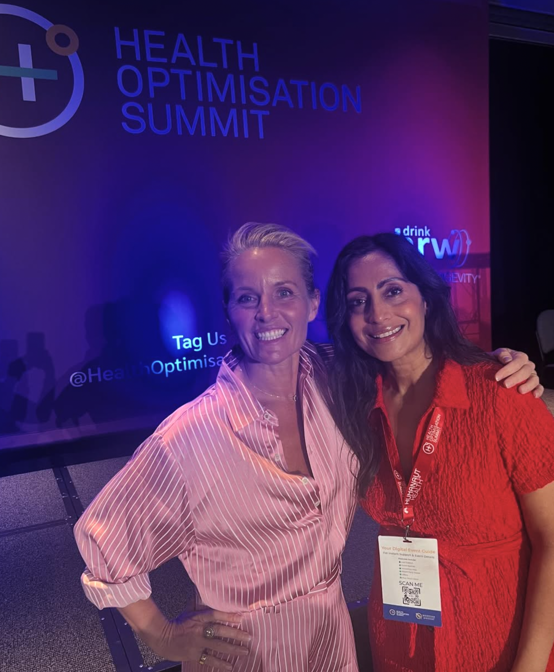 Two women smiling at the Health Optimisation Summit, with a dark background and purple lighting, one in a pink striped shirt and the other in a red dress, both wearing conference badges.
