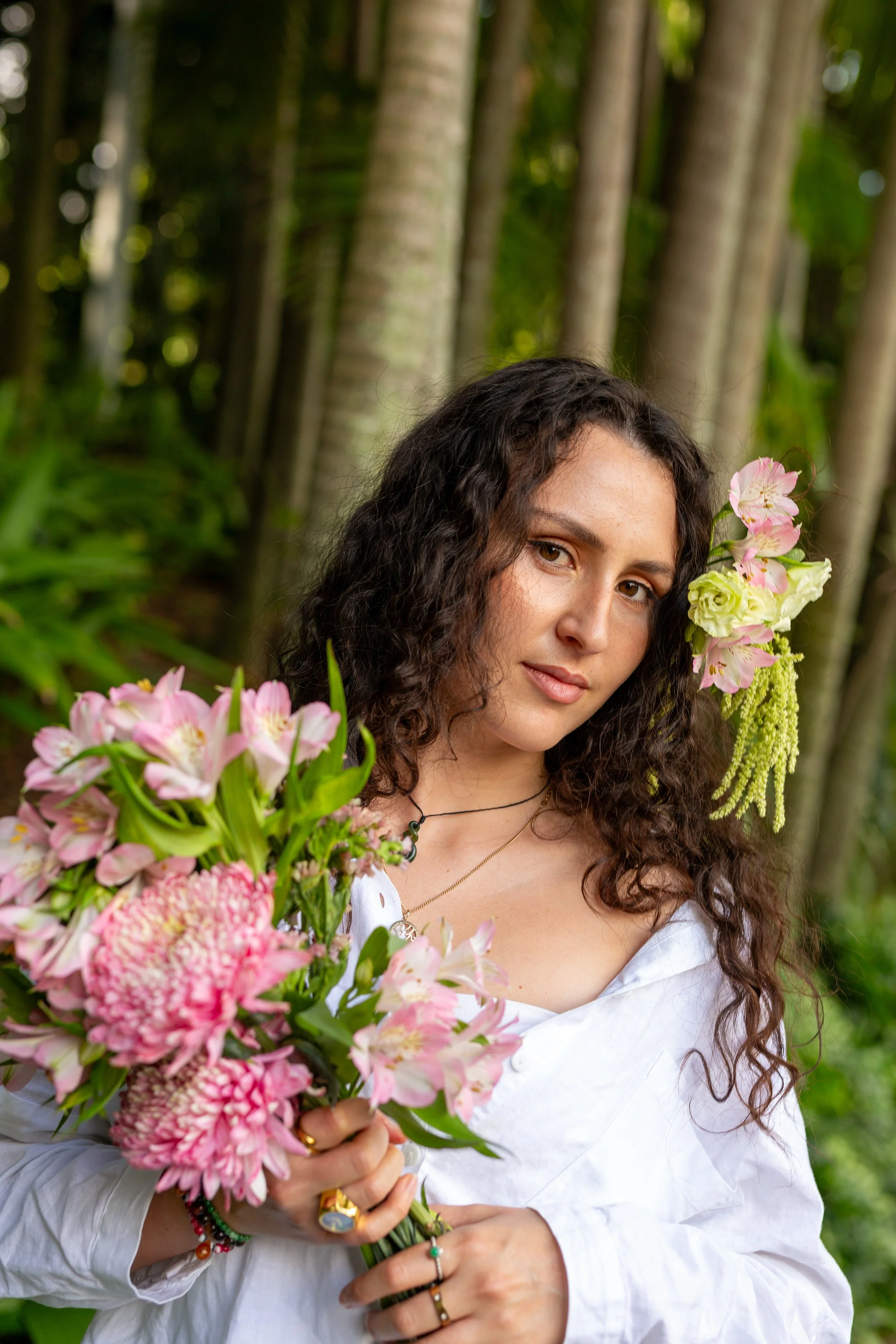 A woman with curly hair holding a large bouquet of pink flowers outdoors, with trees and green foliage in the background.