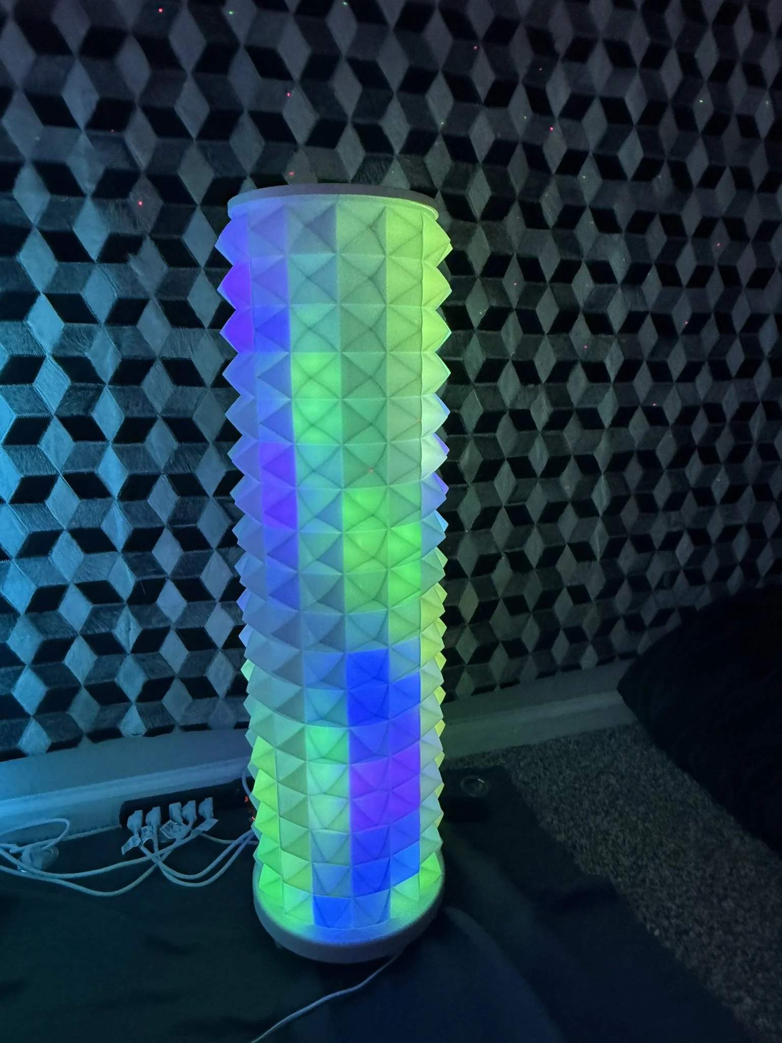 A tall cylindrical lamp with a geometric, diamond-shaped pattern, lit with multicolored LED lights. The background features a hexagonal-tile wall, and there is a power strip with cords nearby.