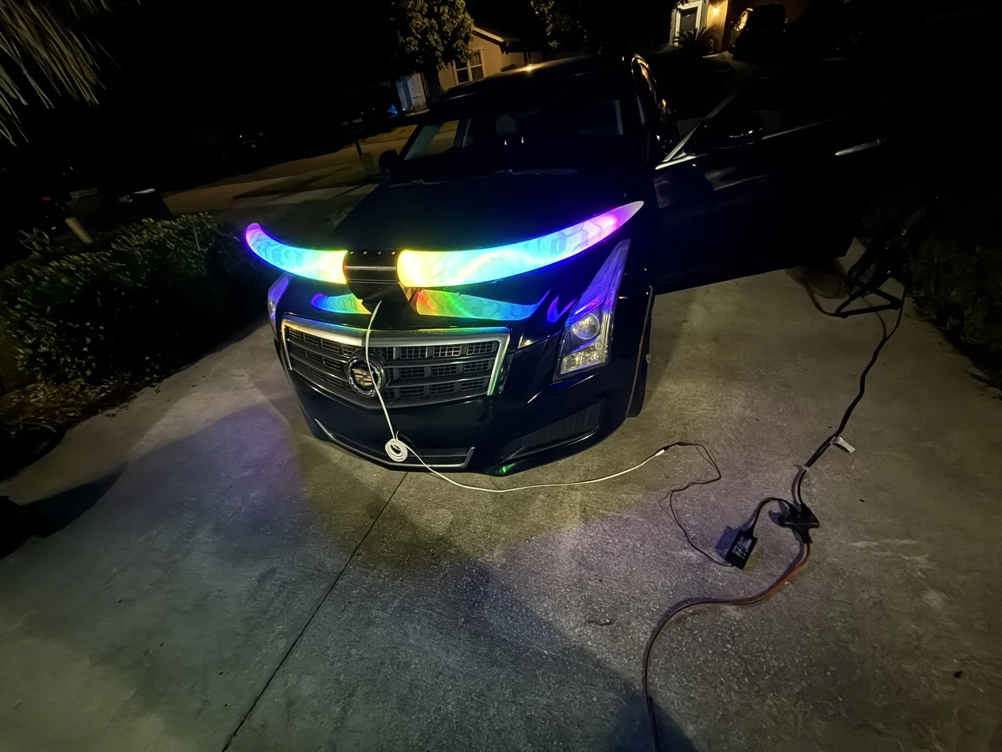 A black car with colorful LED lights on the front is parked on a driveway at night. There are power cords and a power adapter on the ground nearby. The background shows a residential street with houses.