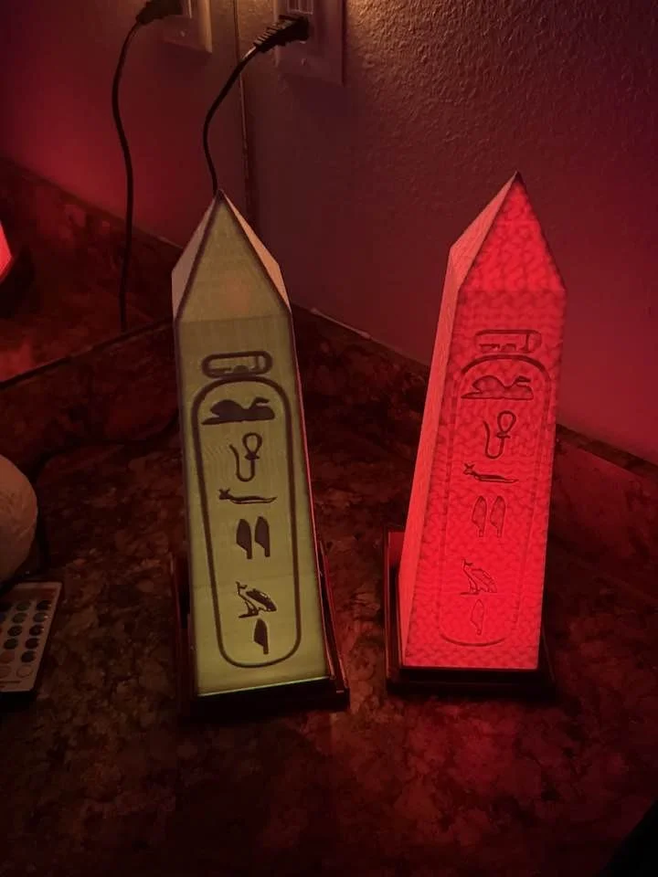Two Egyptian-style pyramid lamps, one emitting yellow light and the other red light, placed on a dark countertop near an electrical outlet.