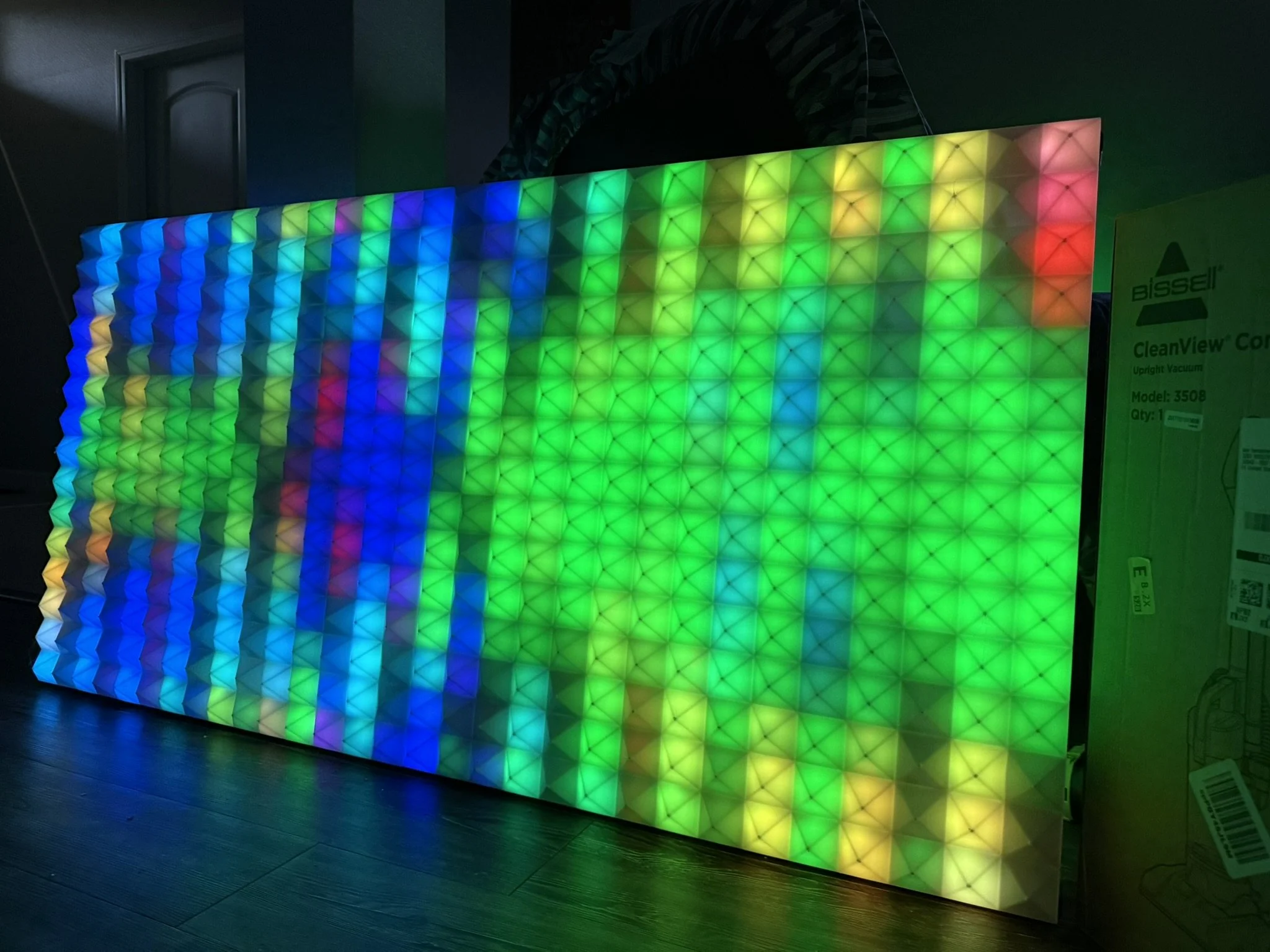 Colorful LED light panel with a geometric pattern showing shades of blue, green, yellow, and red, placed on a dark wooden floor.