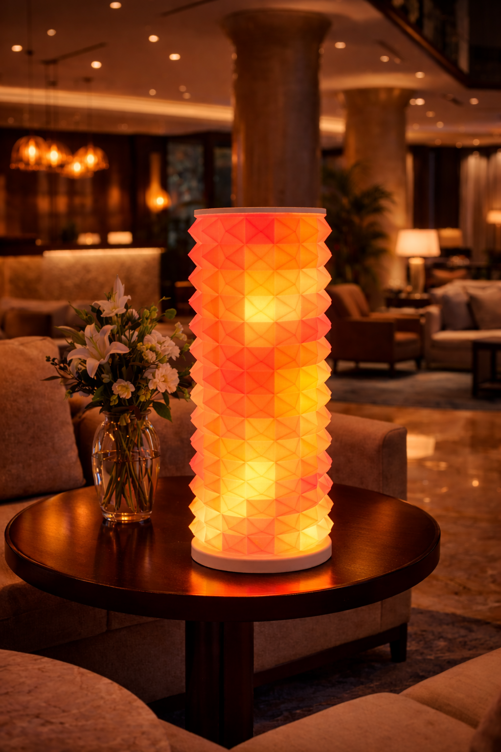A tall, cylindrical, geometric-patterned lamp emitting warm orange and yellow light, placed on a round wooden table next to a vase of white flowers.