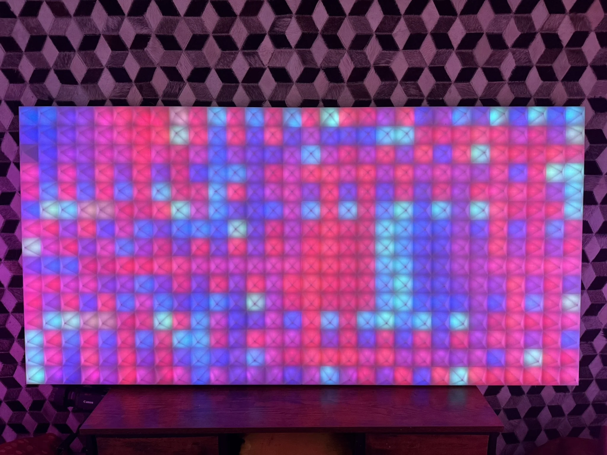 A grid of colorful LED lights displaying pink, purple, blue, and green hues, mounted on a wall with a geometric pattern, with a wooden surface below.