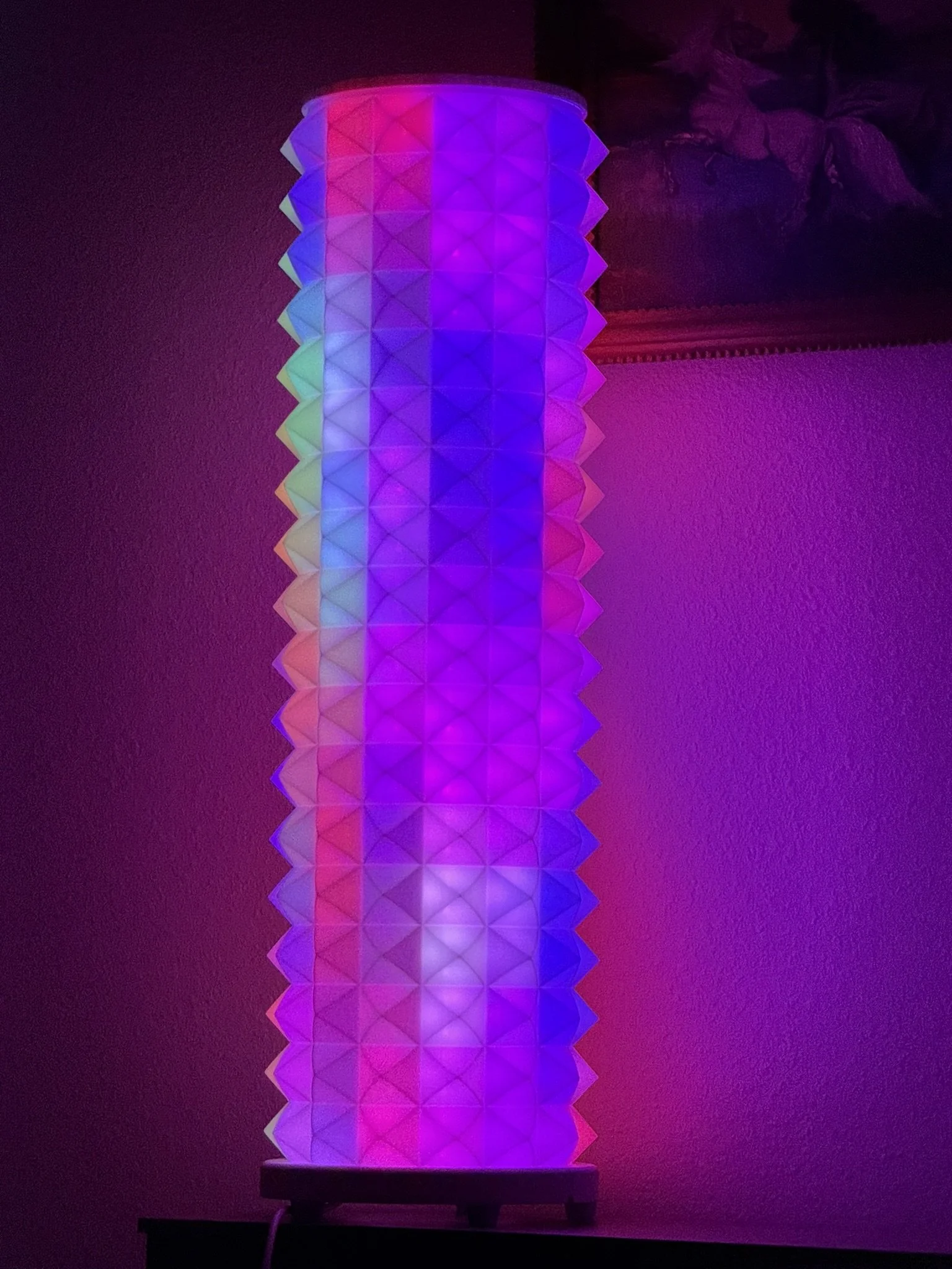 A colorful, geometric-patterned lamp emitting shades of purple, pink, blue, and green, placed on a dark surface against a purple-lit background.