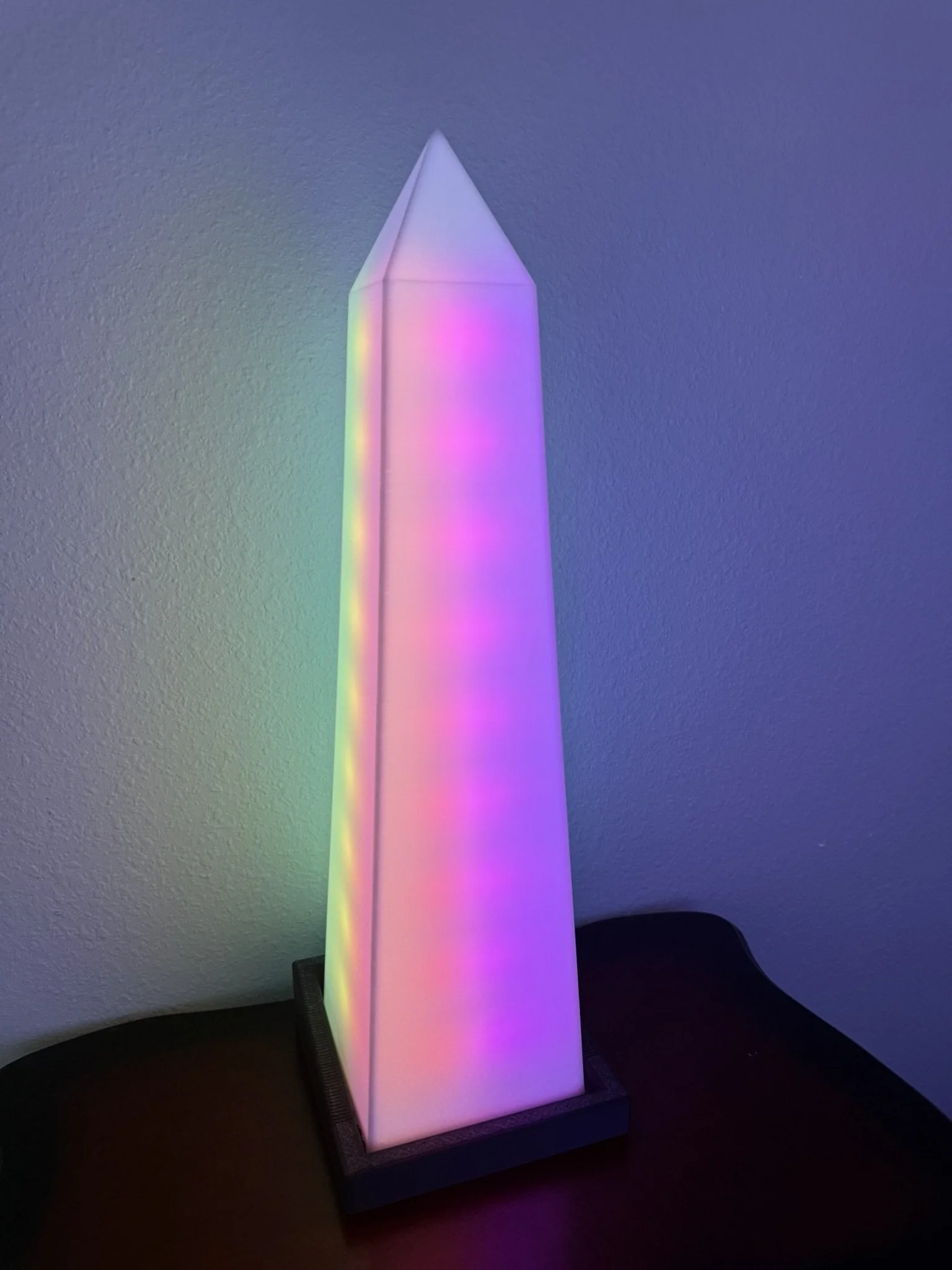 A tall, rectangular LED lamp emitting multicolored light, placed on a dark table against a plain wall.