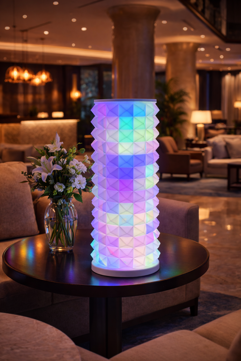 A colorful geometric illuminated lamp on a round table with a vase of flowers in a cozy lounge area.