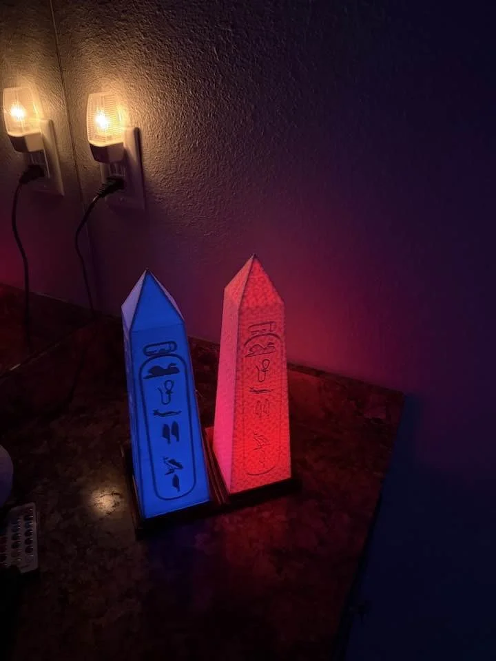 Color-changing pyramid-shaped lamps with mathematical equations on the sides, placed on a dark countertop near wall outlets with night lights.