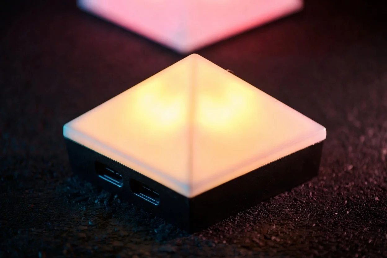 A square device with a glowing, colorful illuminated top surface and two USB ports on the side, placed on a dark textured surface.